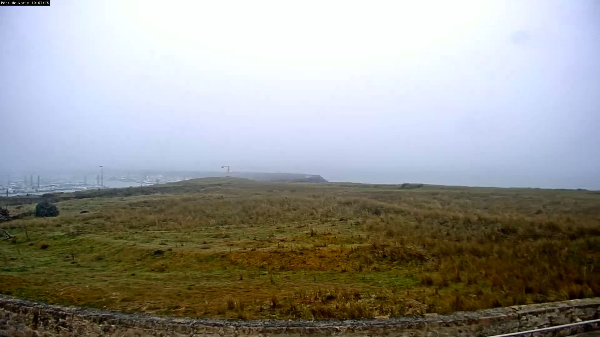 A foggy coastal landscape shows a grassy expanse leading to a hazy ocean, with a distant port visible in the background.