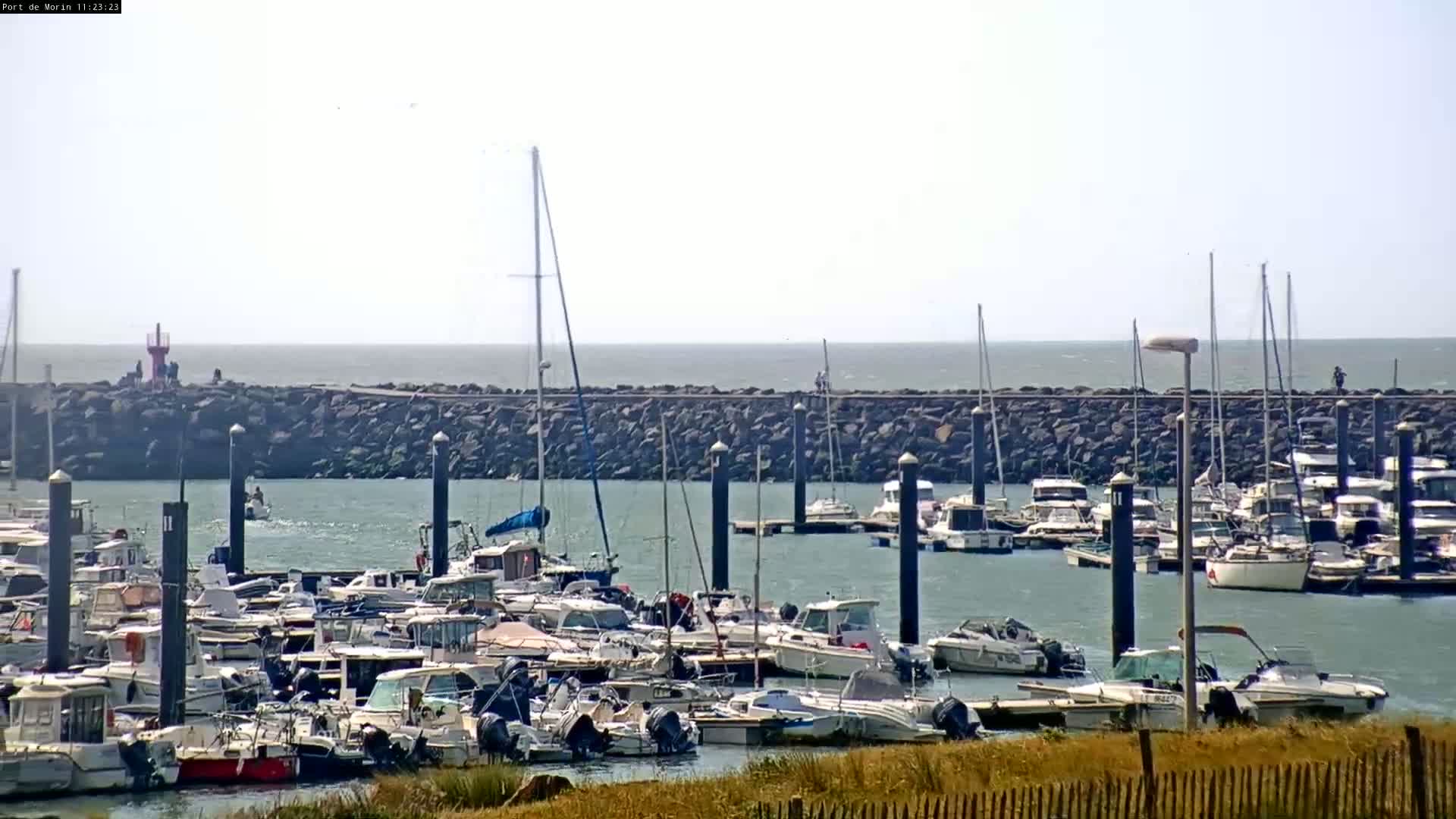 A marina filled with numerous boats is nestled behind a rocky breakwater, overlooking a calm sea under a bright, mostly clear sky.