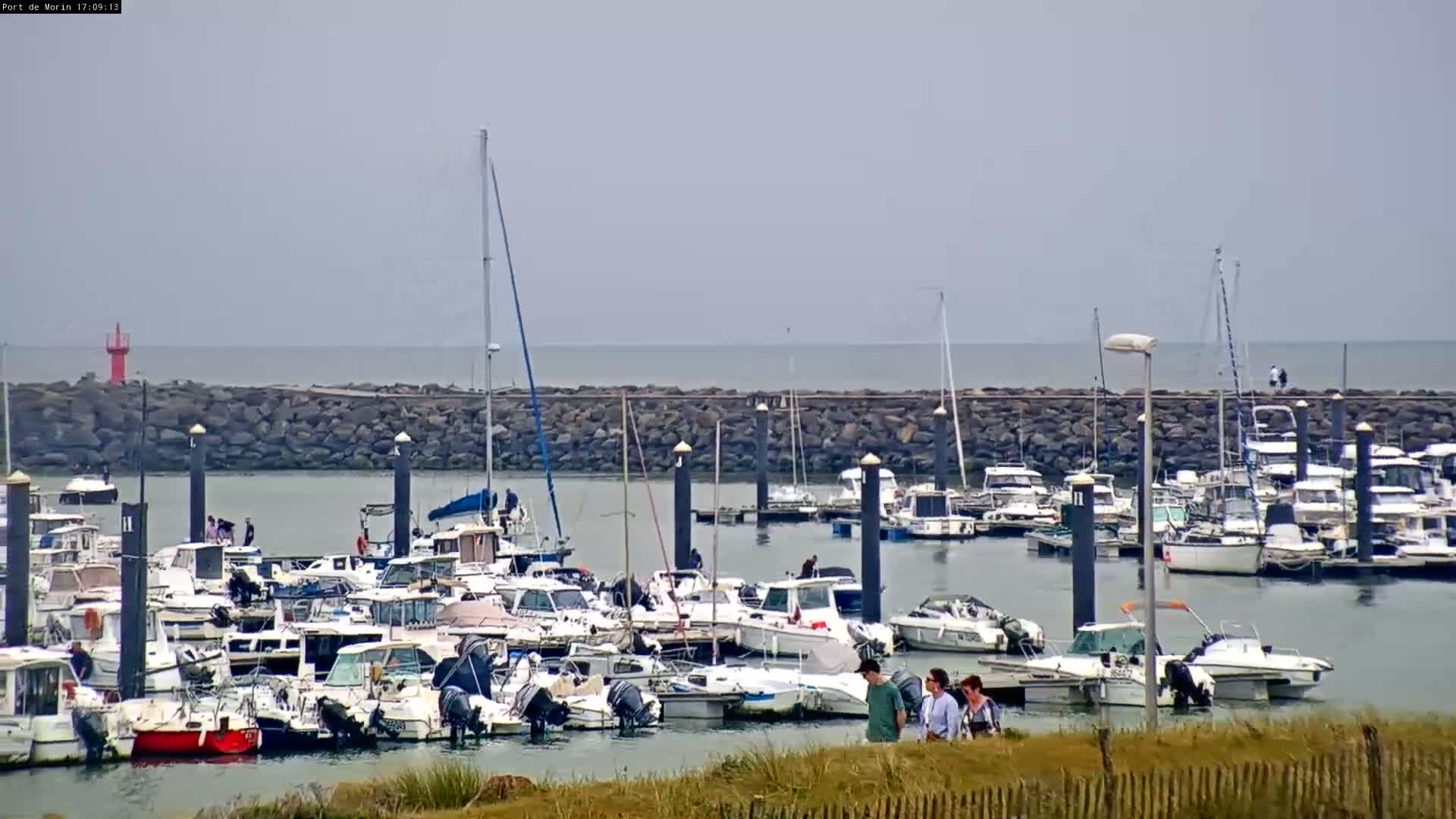 A calm harbor filled with numerous boats and a few people walking along the shore under an overcast sky.