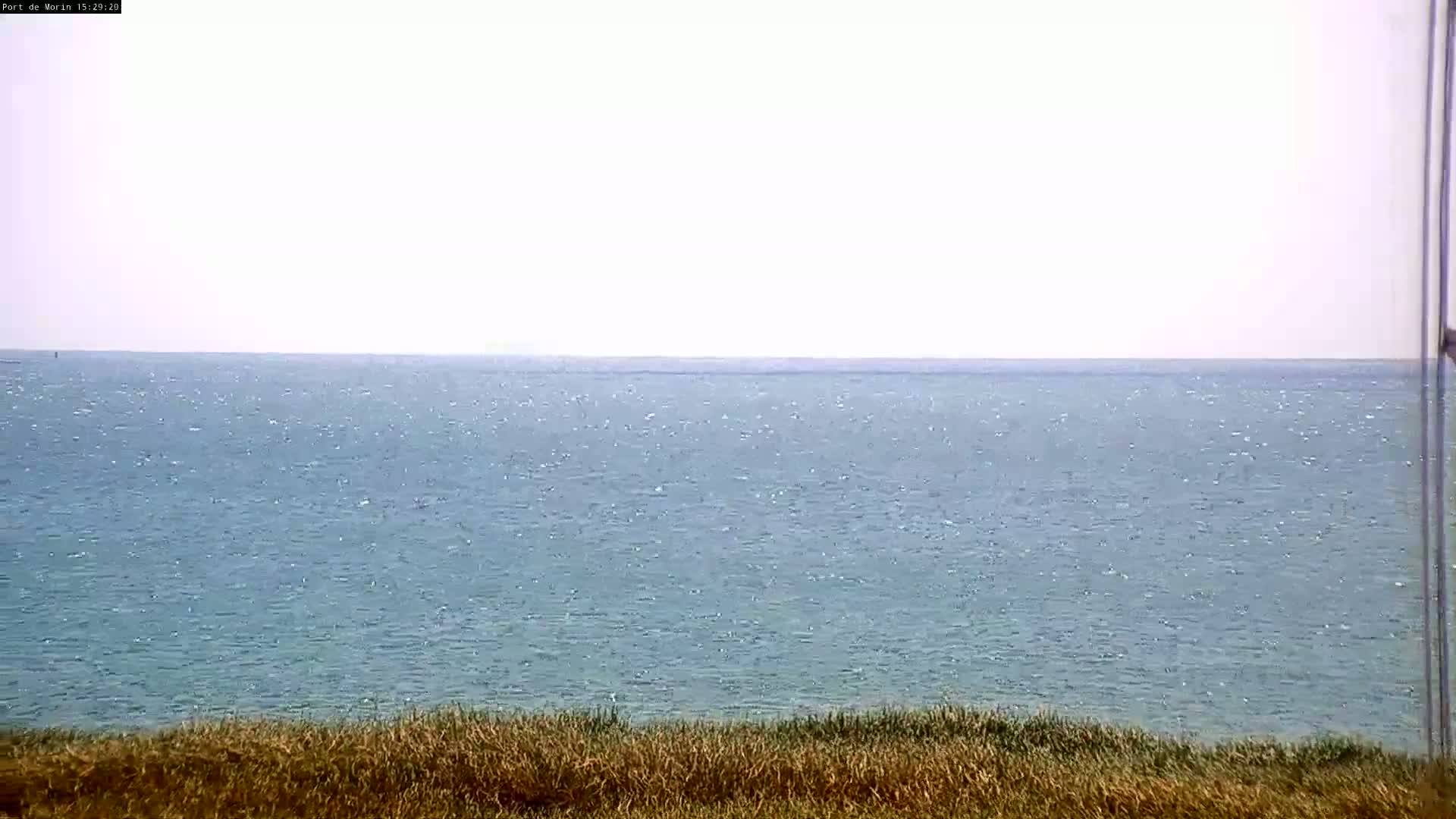 A calm, light-blue ocean stretches to a hazy horizon from a vantage point of dry, tan grass.