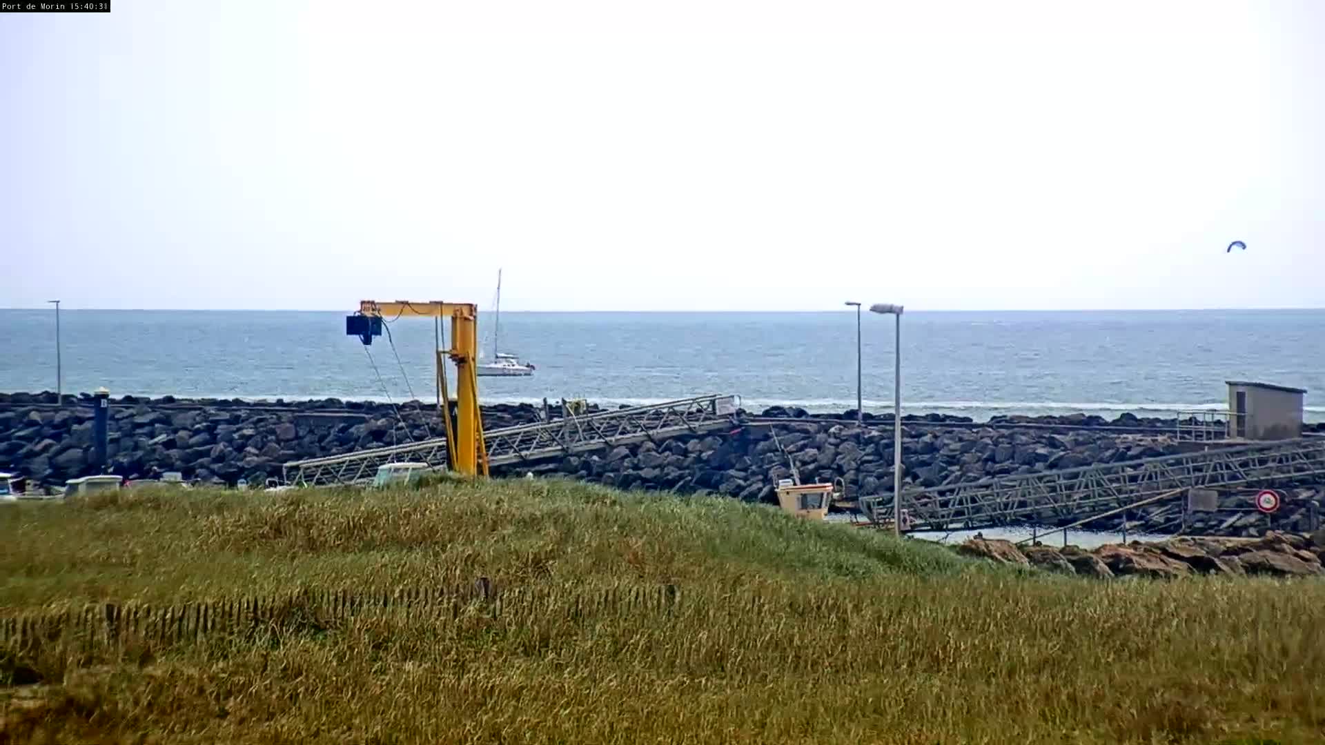 A yellow crane sits on a rocky shore next to a calm ocean with a sailboat and a kitesurfer visible in the distance, under an overcast sky.