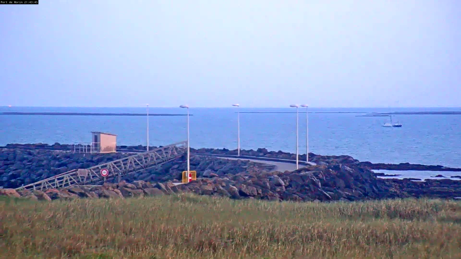 A rocky shoreline with a small building and a metal walkway extends into a calm, blue sea under a clear sky.