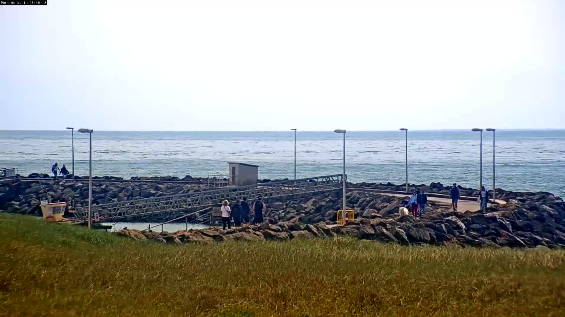 A rocky shoreline with a small metal footbridge and several people walking, overlooking a calm ocean under a mostly cloudy sky.