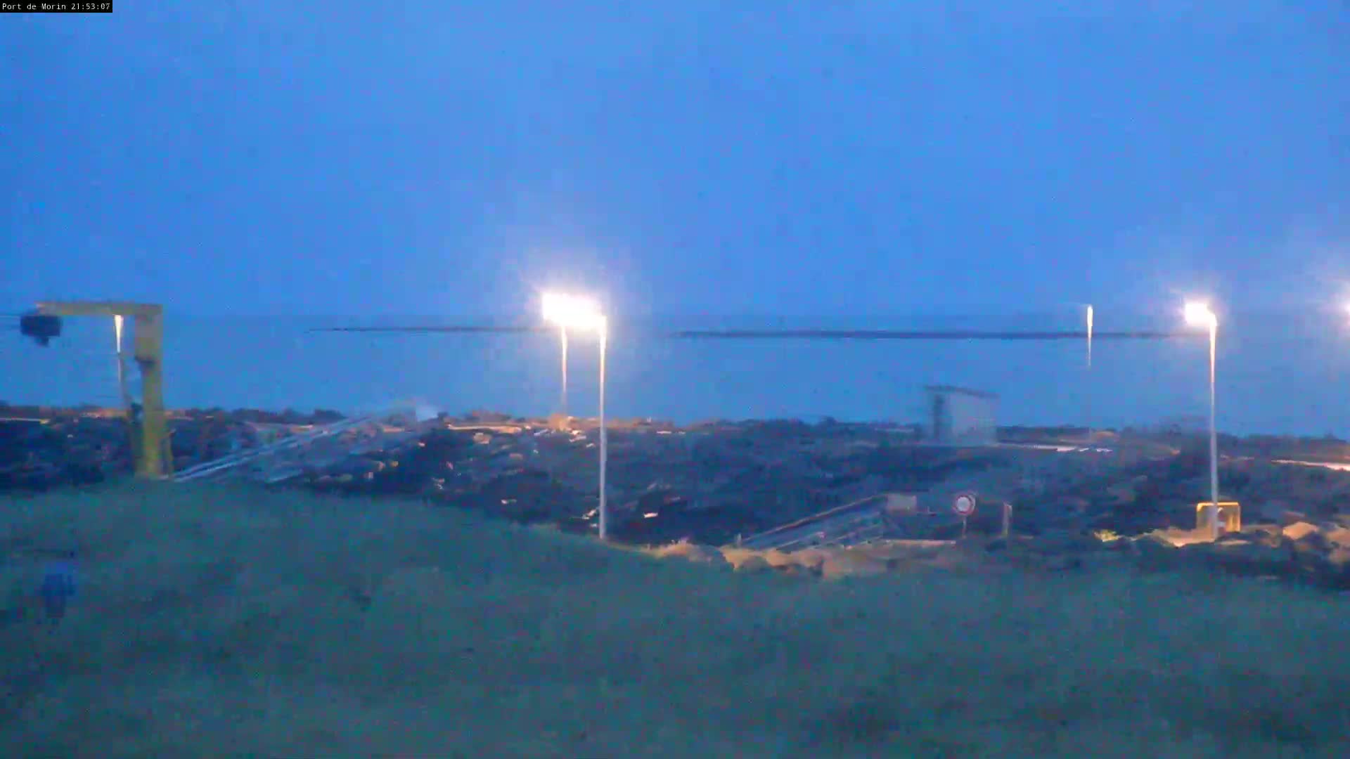 A dark, mostly clear twilight sky overlooks a rocky shoreline with a few lit lamp posts and a small structure near the water.