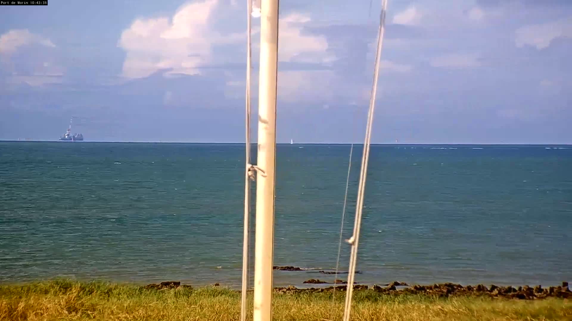 A partly cloudy sky overlooks a calm, teal ocean with a distant ship and sailboat, viewed from behind a tall, light-colored pole.