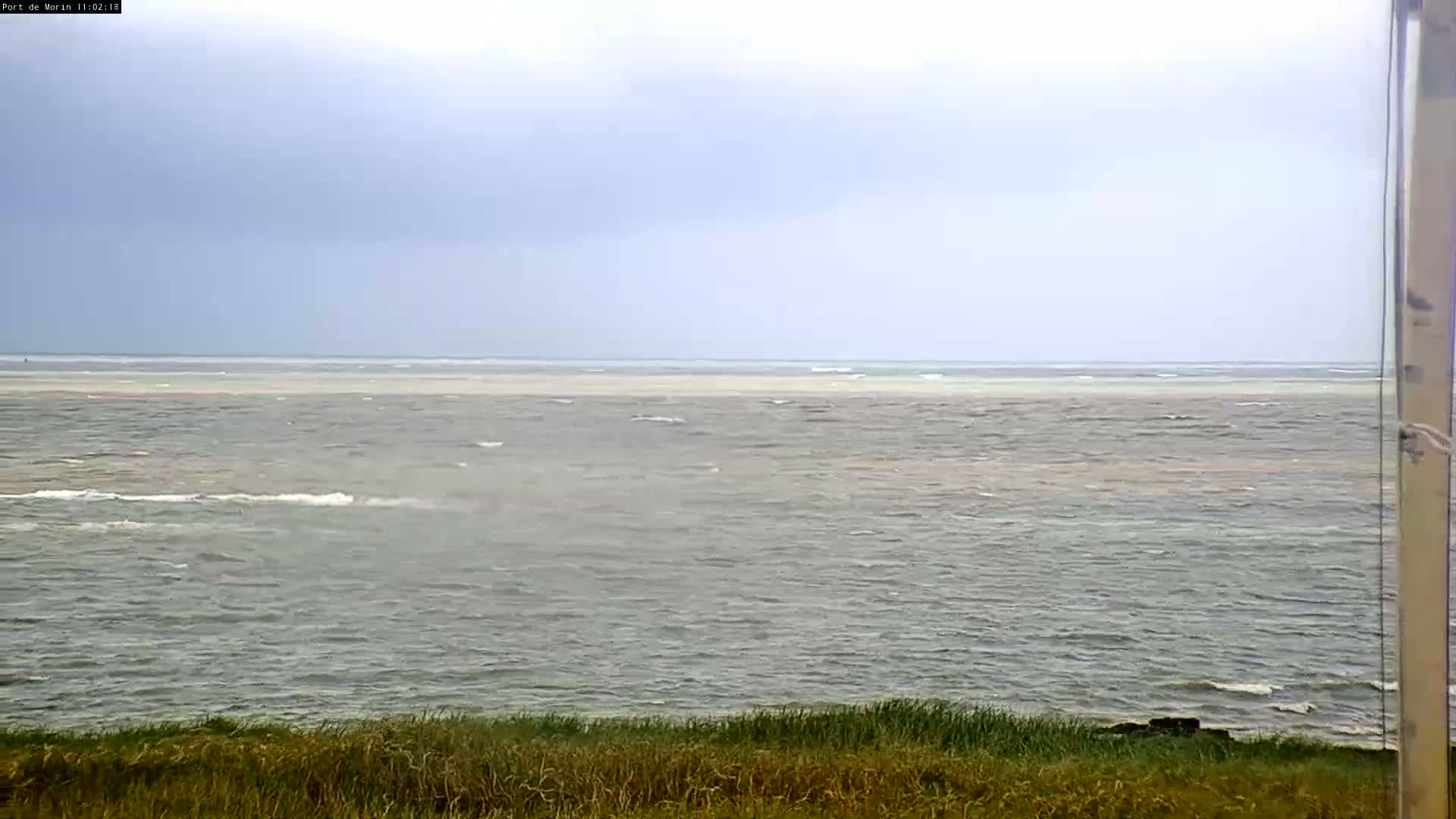 A slightly overcast sky overlooks a choppy grey-green sea with a lighter colored band near the horizon, bordered by grassy dunes in the foreground.