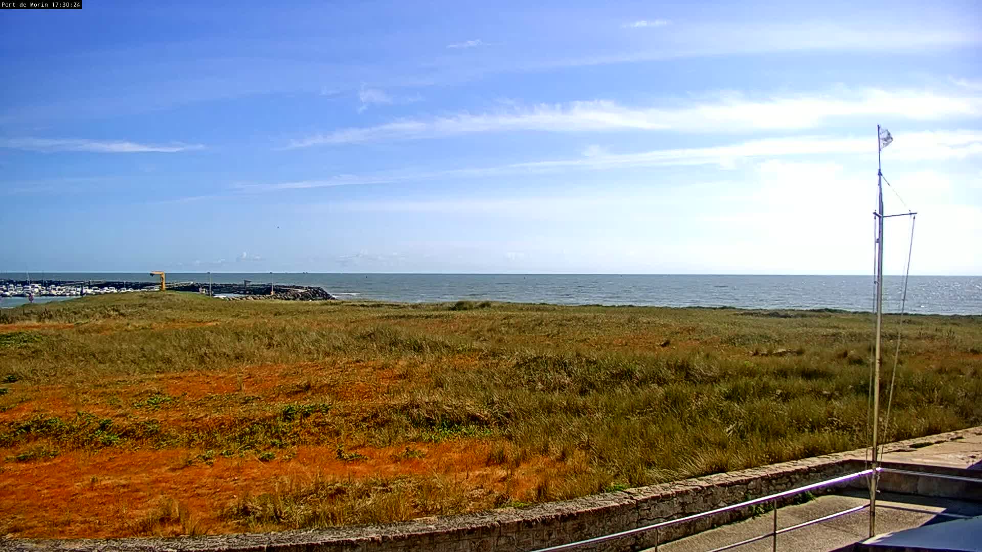 A mostly sunny day reveals a calm ocean stretching to the horizon behind a field of dried, orange-tinged grass, a small harbor visible in the distance.