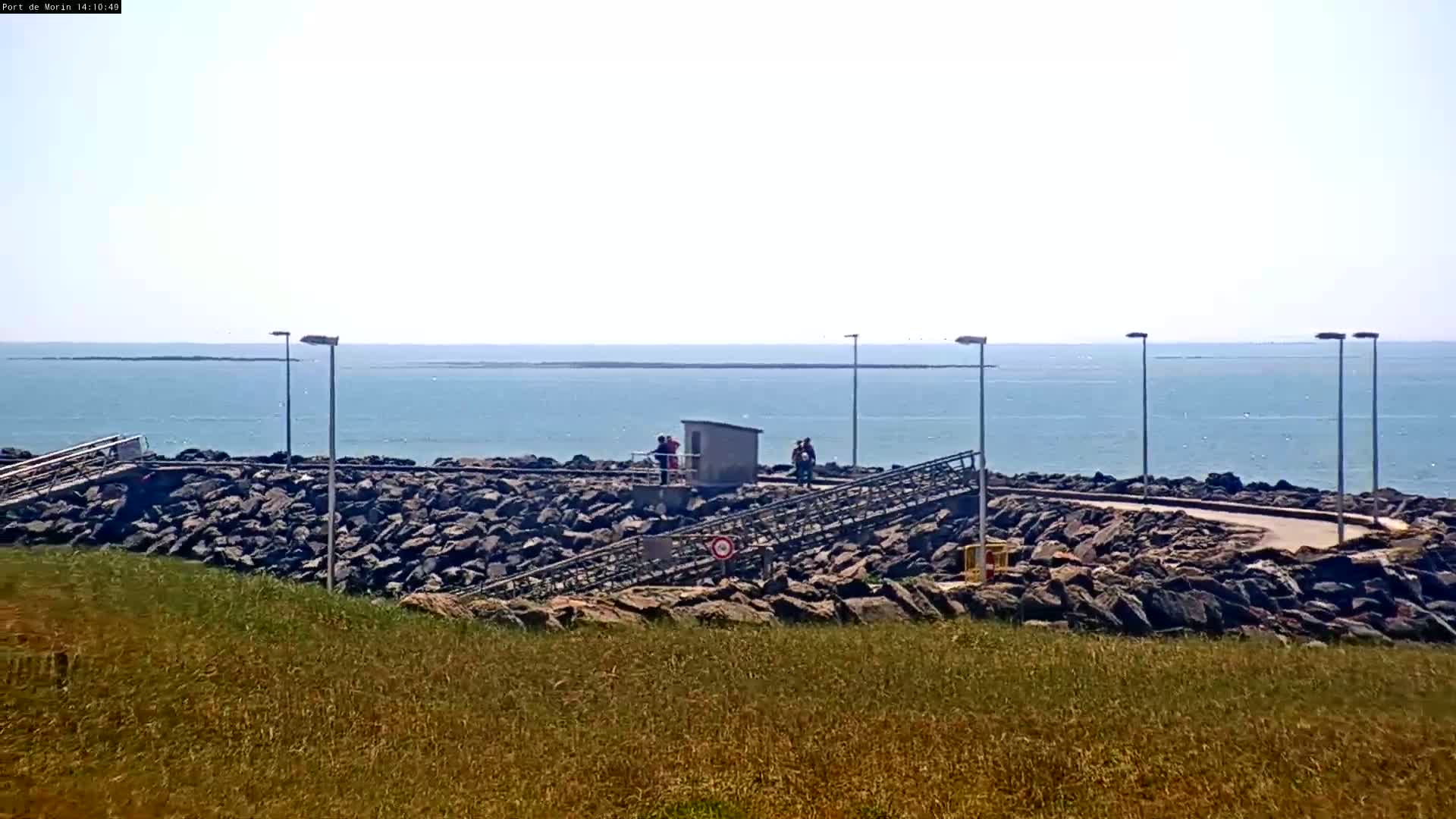 A rocky shoreline with a few people standing near a small building overlooks a calm, blue ocean under a bright, mostly clear sky.