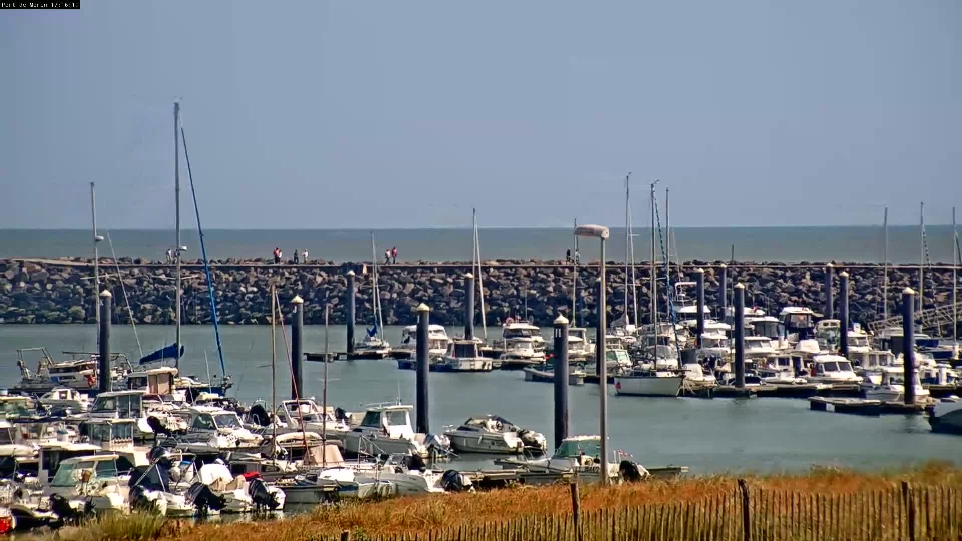 A marina filled with numerous boats is situated behind a rock wall, with people walking along the wall under a clear, sunny sky.