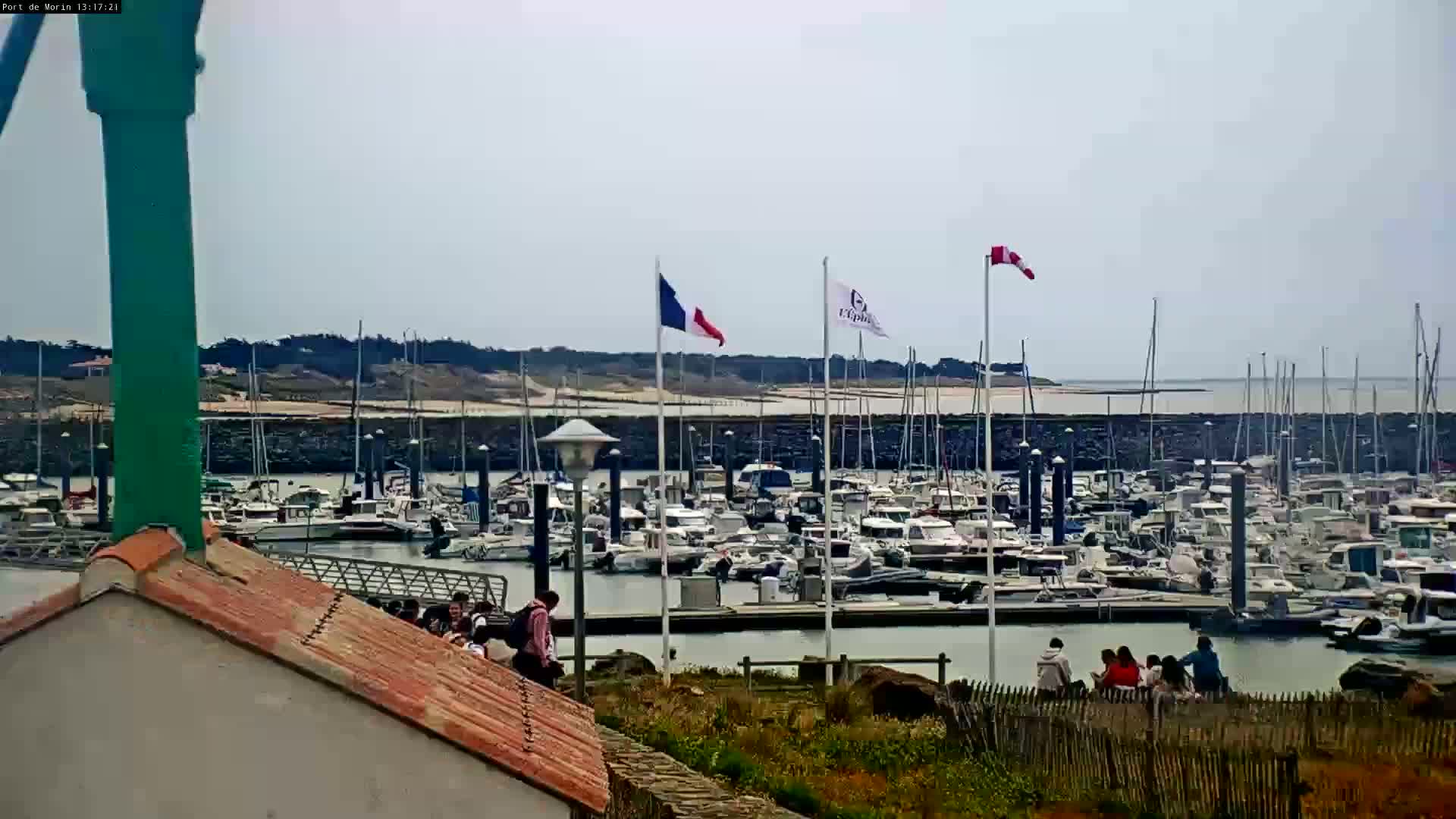 A marina filled with numerous boats is seen on a partly sunny day, with a few people gathered near a low stone wall overlooking the water.