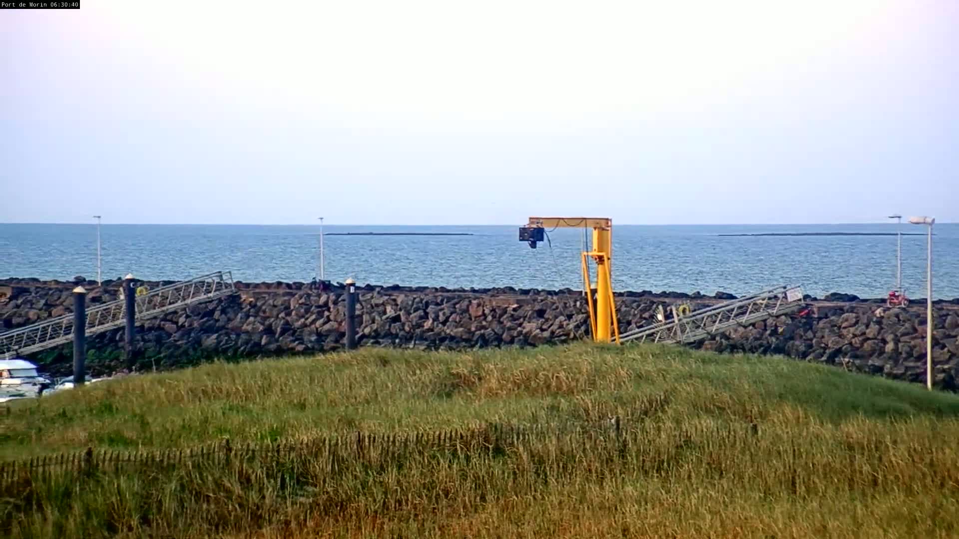 A yellow crane stands near a rocky shoreline overlooking a calm ocean under a clear sky.