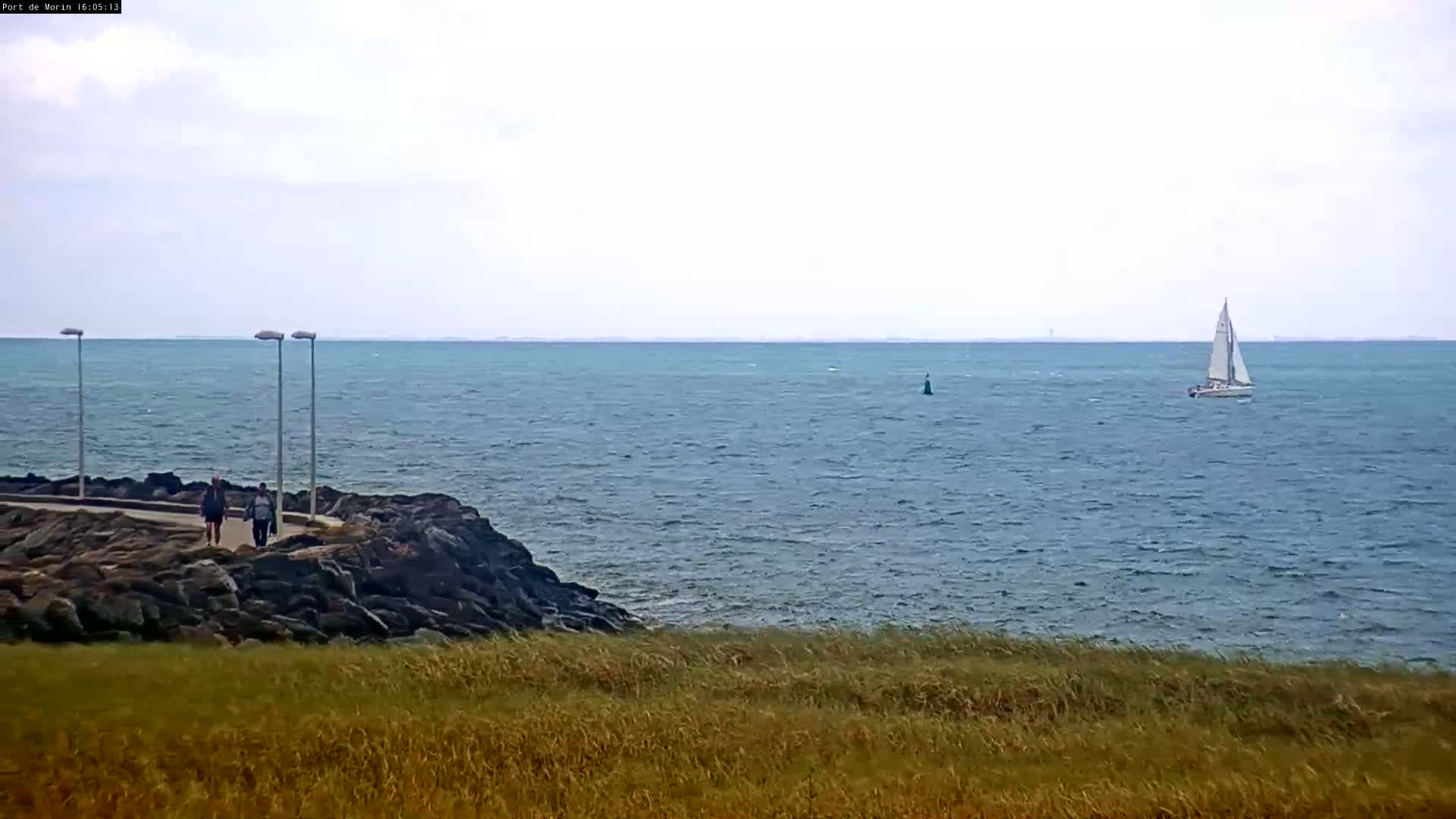 A sailboat is visible on a somewhat choppy sea, near a rocky shoreline with two people walking along a path, under a cloudy sky.