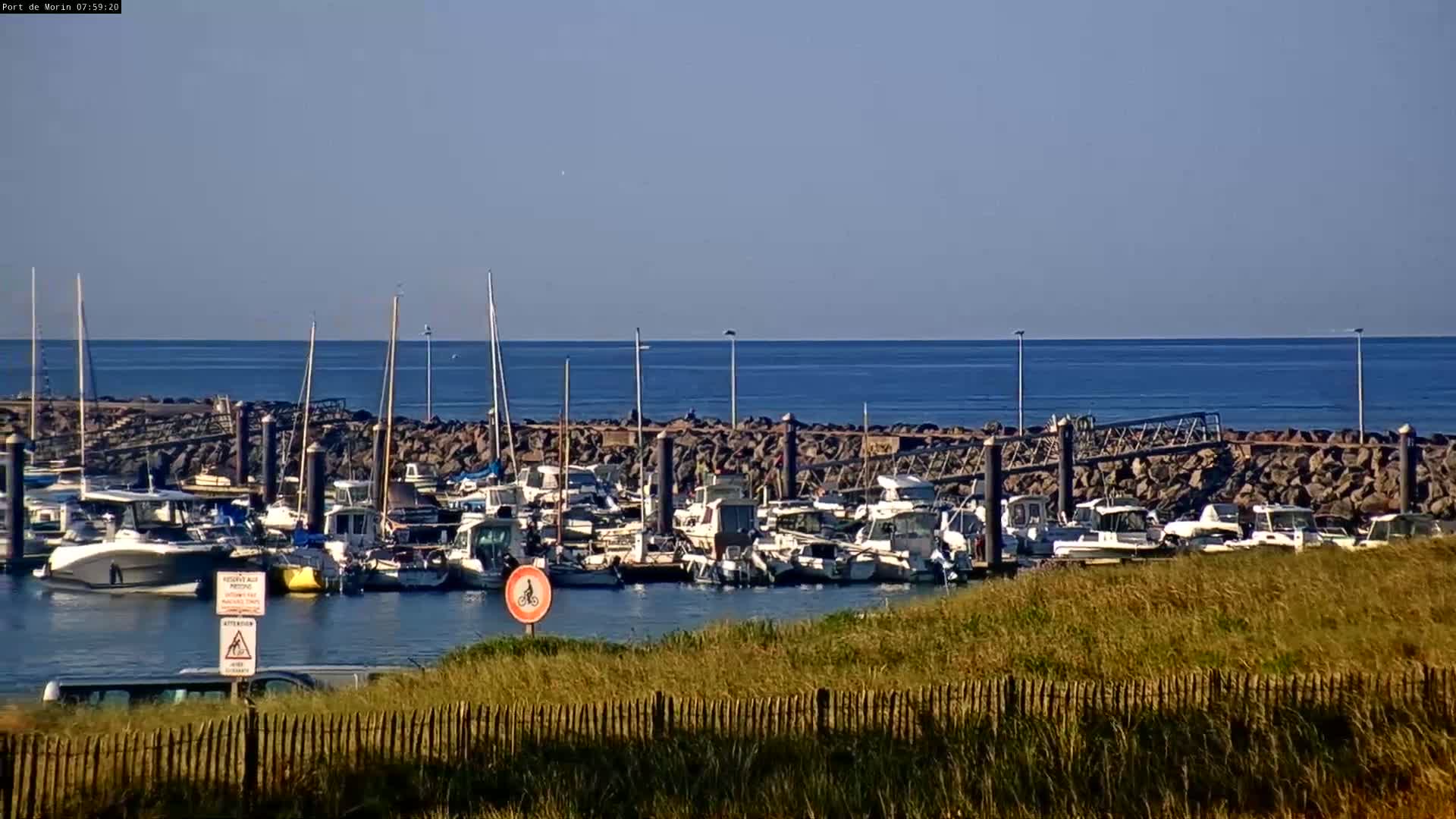 Many boats are docked in a harbor protected by a rock wall, with a calm blue sea extending to the horizon under a clear sky.