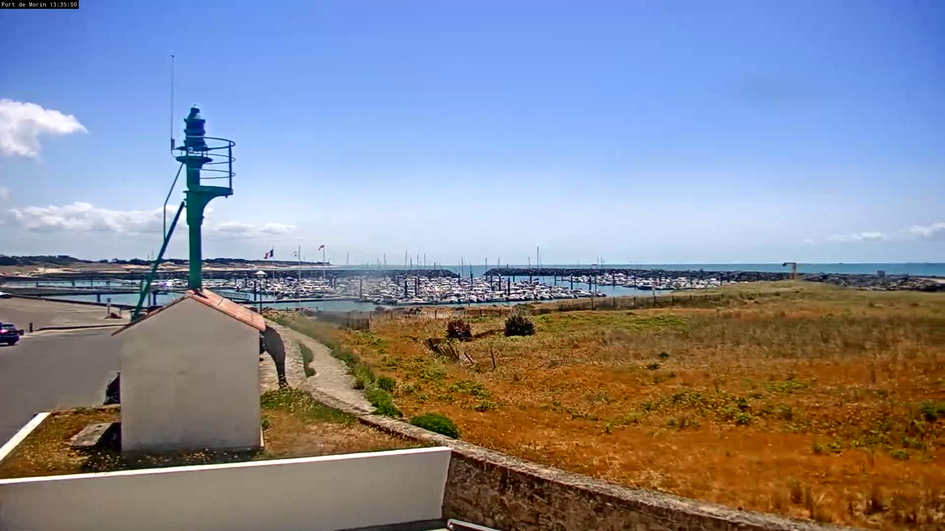 A green signal light stands near a small white building overlooking a marina filled with boats on a sunny day with a mostly clear blue sky.