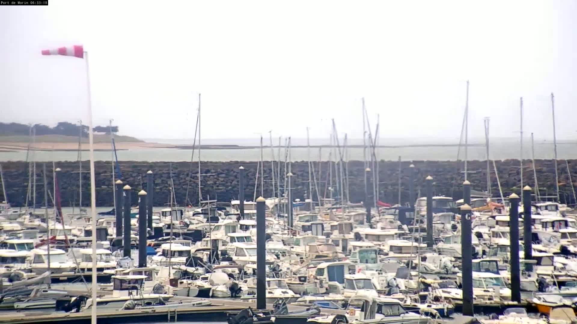A marina filled with numerous boats and yachts is situated near a calm body of water under an overcast sky, with a windsock indicating a light breeze.