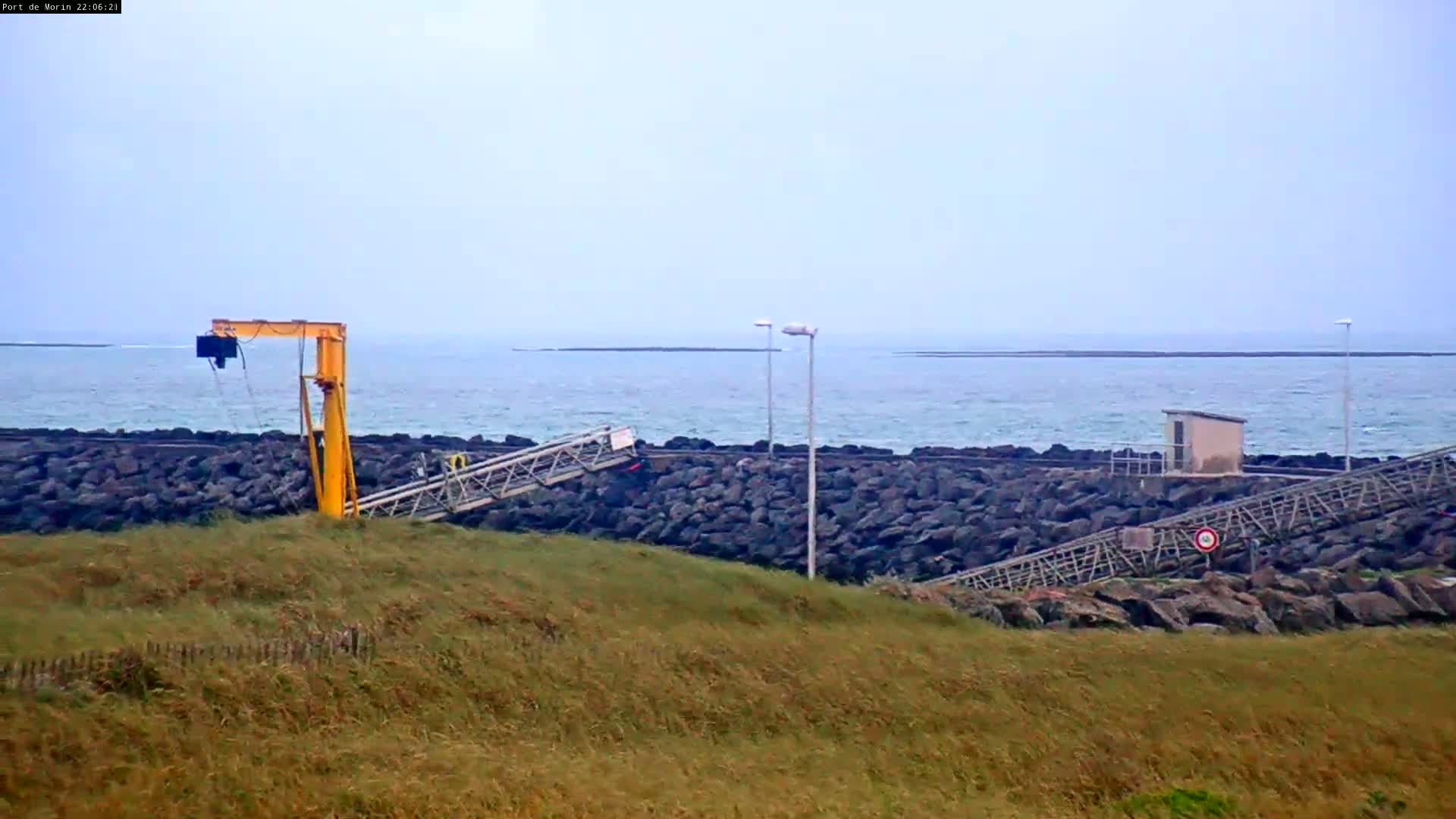 A yellow crane and a metal walkway are situated on a rocky shore next to a calm sea under an overcast sky.