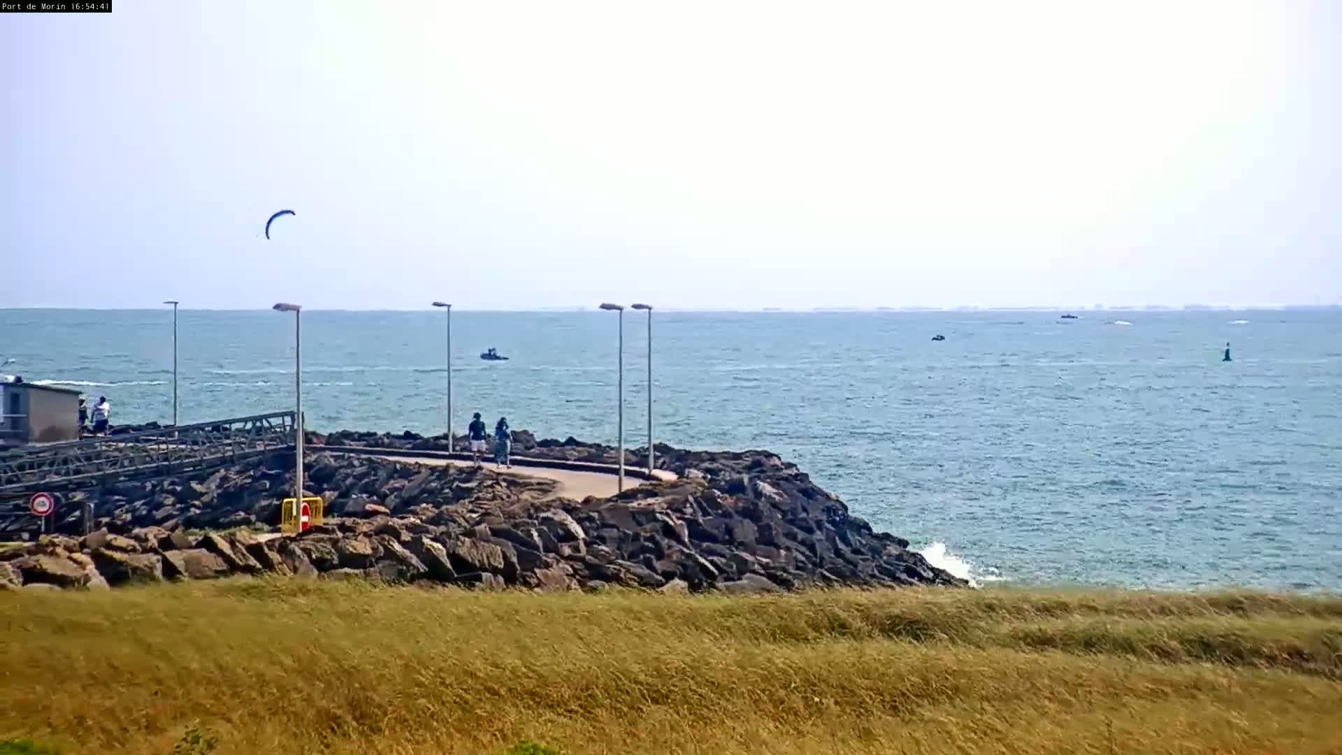 A rocky shoreline with a paved walkway overlooks a calm sea on a sunny day, with a kite surfer visible in the distance.