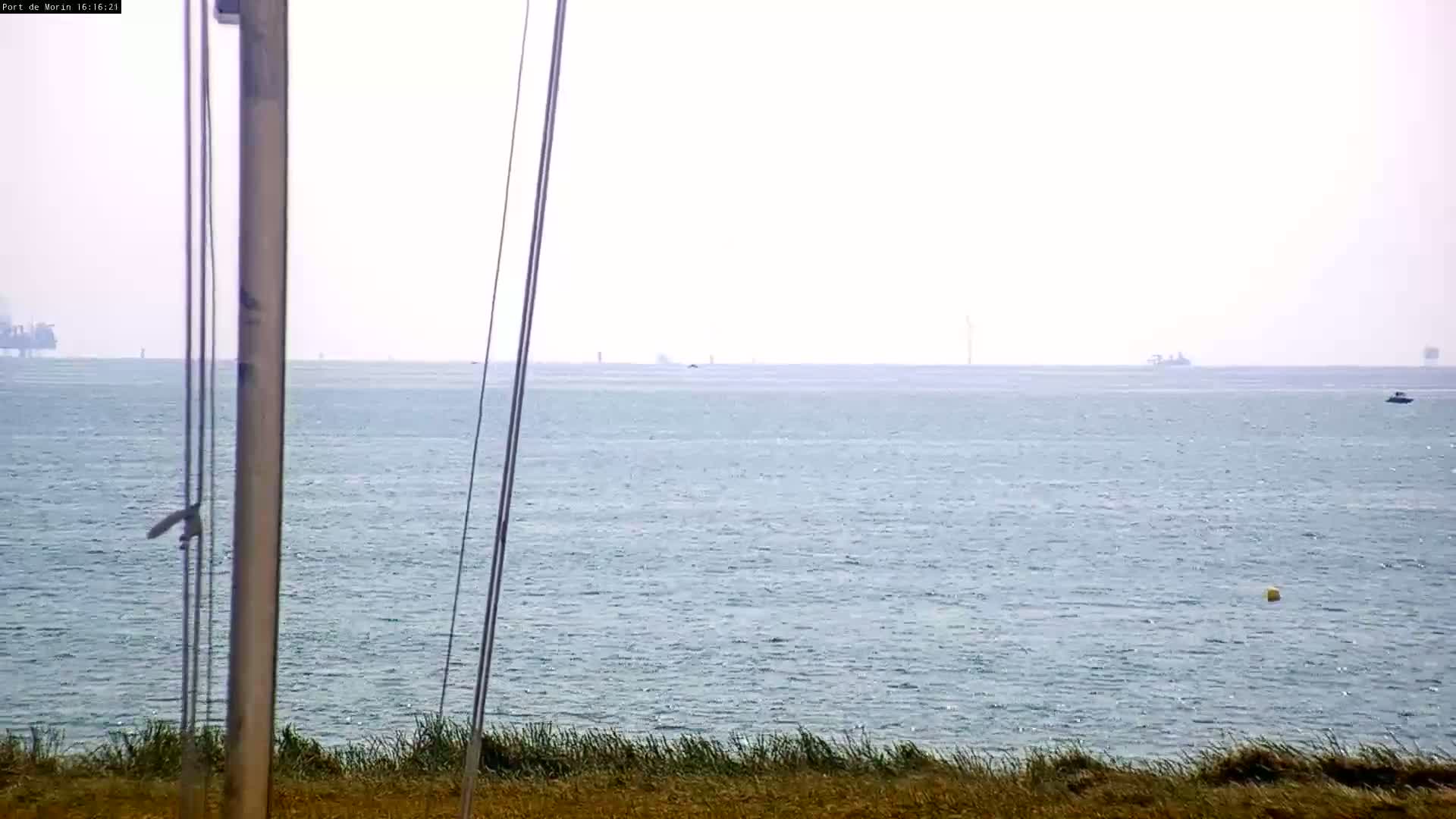 A calm, hazy ocean is seen from a coastal vantage point, with distant ships and wind turbines visible on the horizon.
