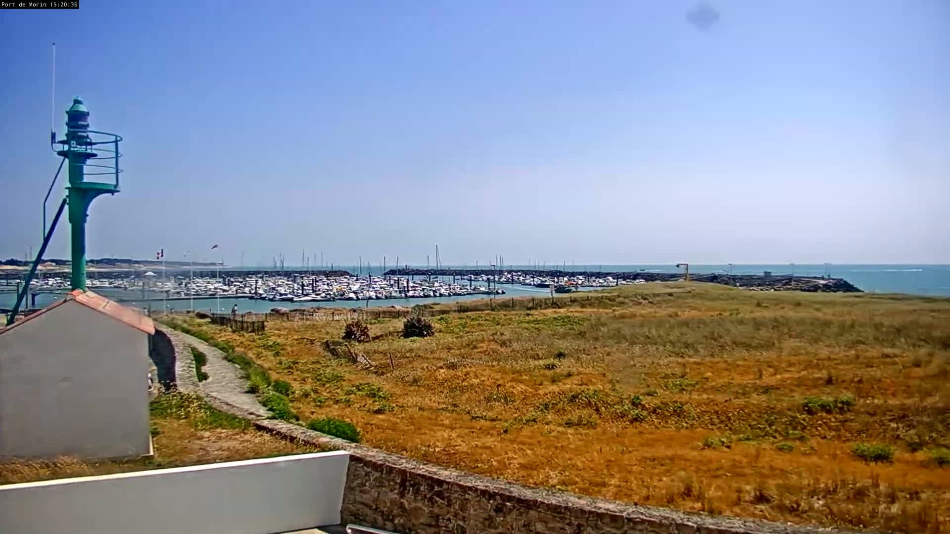 A small green lighthouse stands overlooking a marina filled with numerous boats under a clear blue sky on a sunny day.