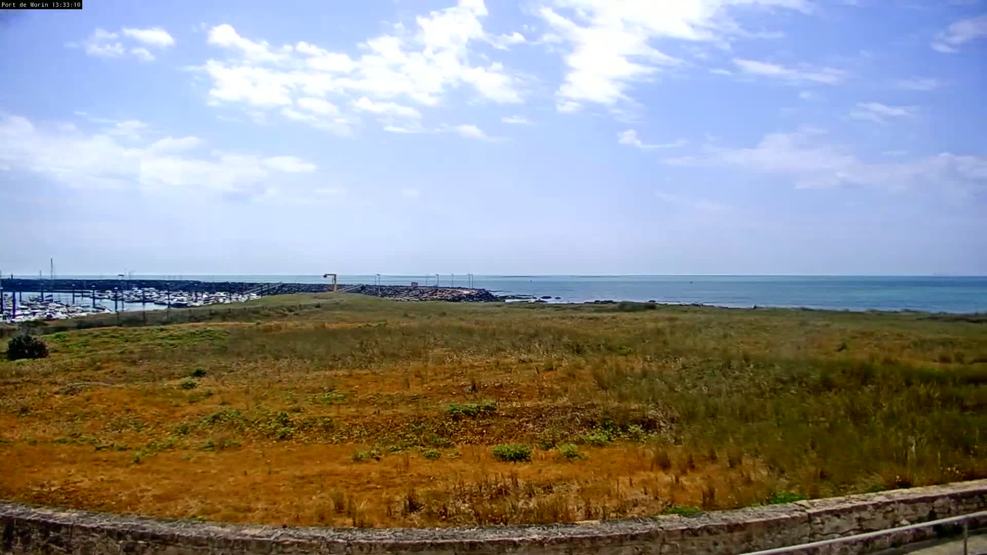 A partly cloudy sky overlooks a dry, grassy field bordering a calm ocean with a marina in the distance.
