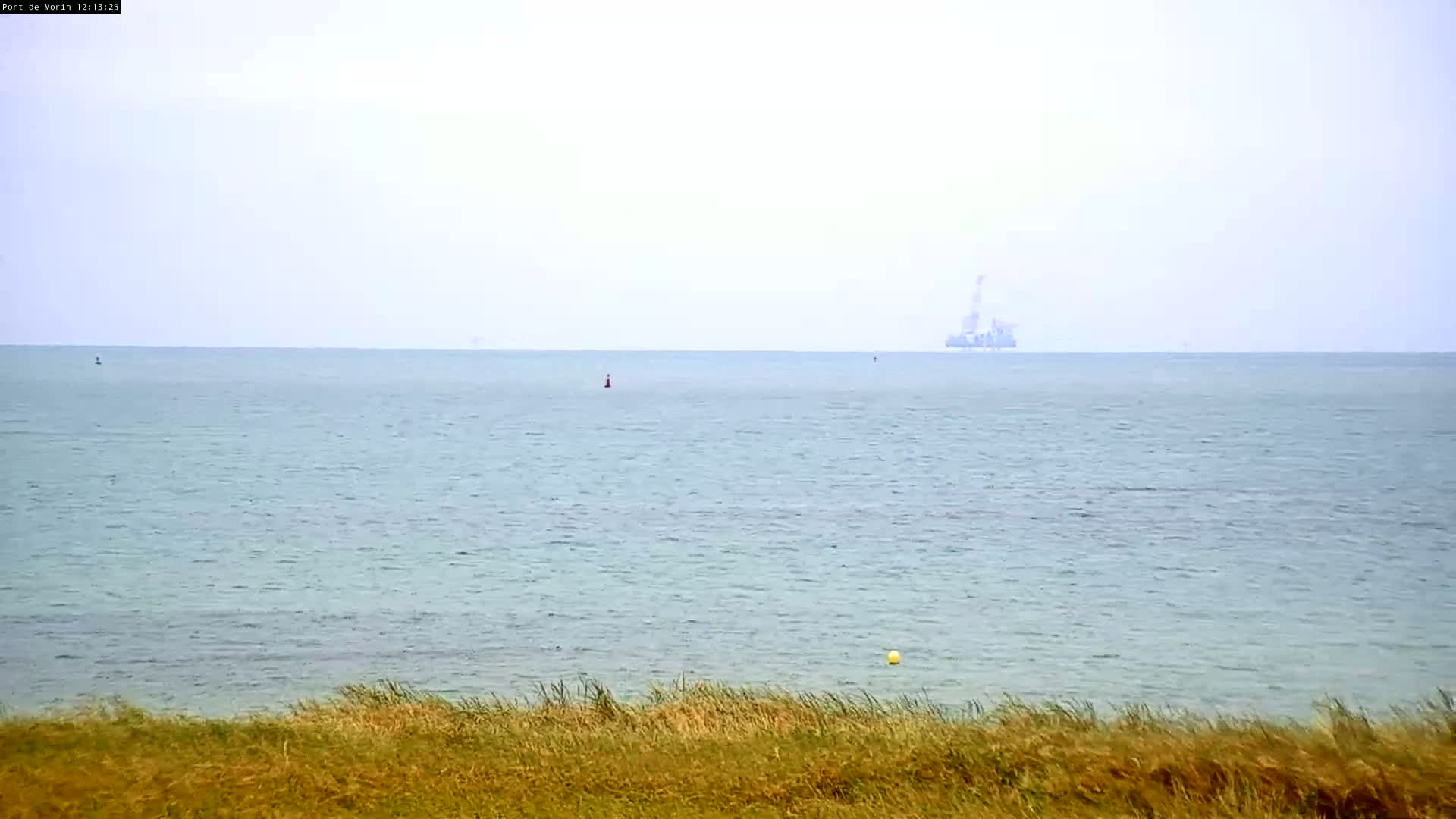 A distant offshore oil rig is visible on a hazy, calm ocean, viewed from a grassy dune.