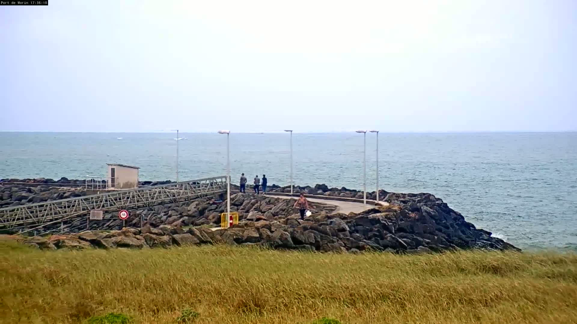 A rocky shoreline with a paved walkway and several people stretches out into a calm sea under an overcast sky.