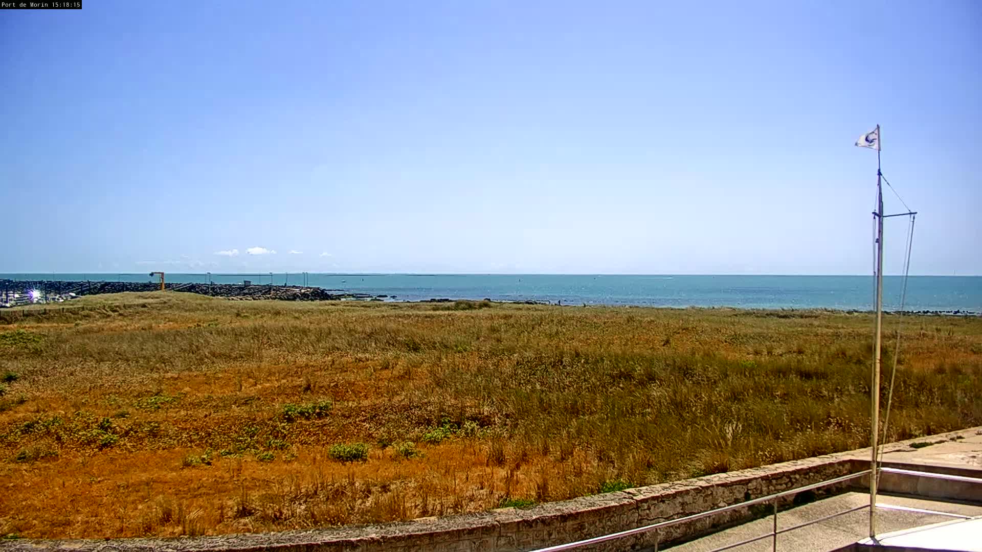 A flat expanse of dry, golden-brown grass stretches between a low stone wall and a calm, teal ocean under a clear blue sky.