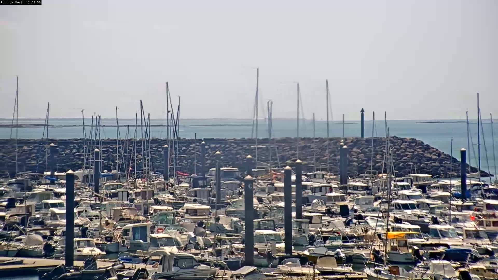 A densely packed marina filled with numerous boats is situated behind a rock breakwater, overlooking a calm, light-blue sea under a hazy sky.