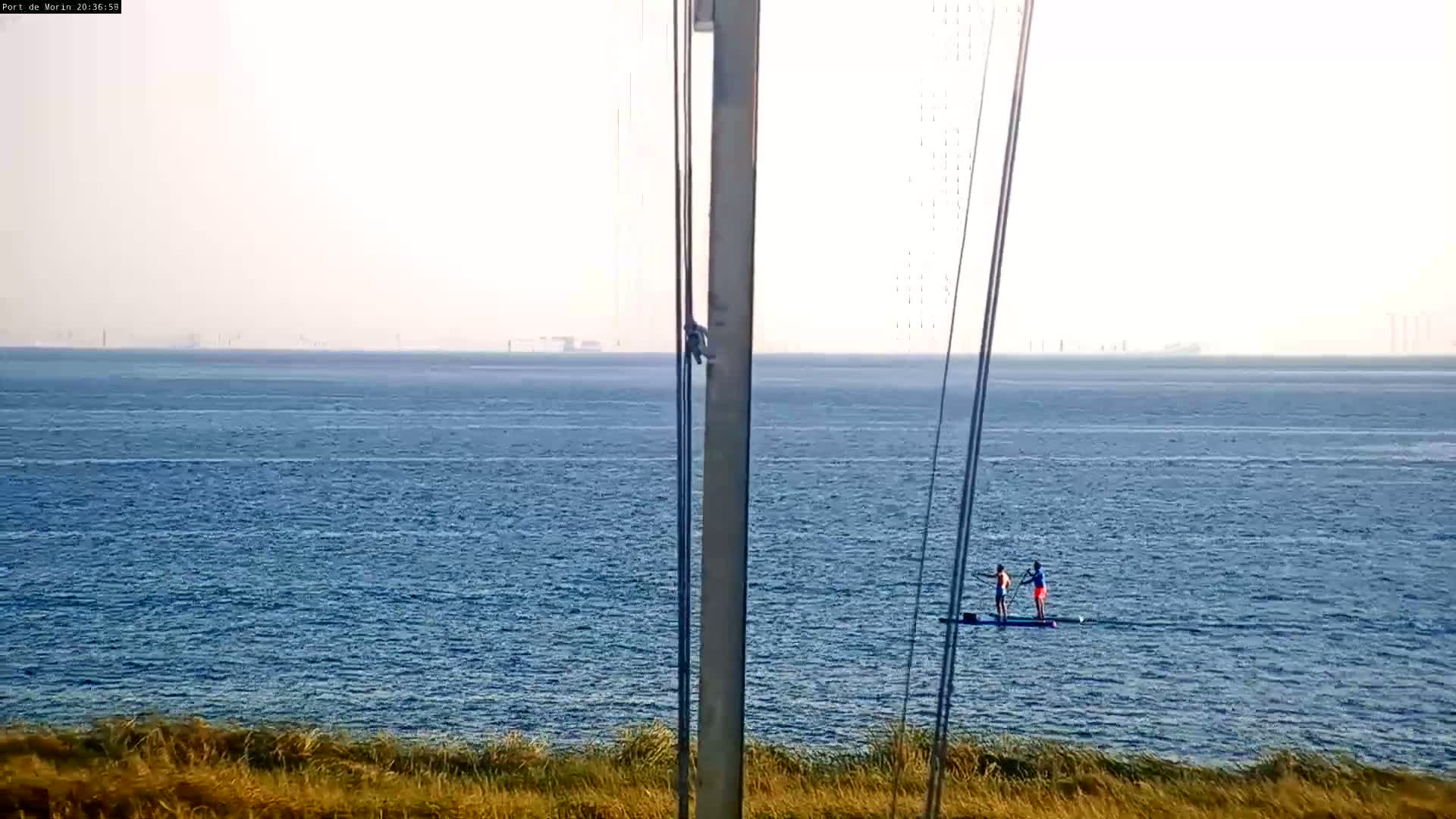 Two people stand on paddleboards in a calm, blue ocean under a hazy sky, with distant land visible on the horizon.