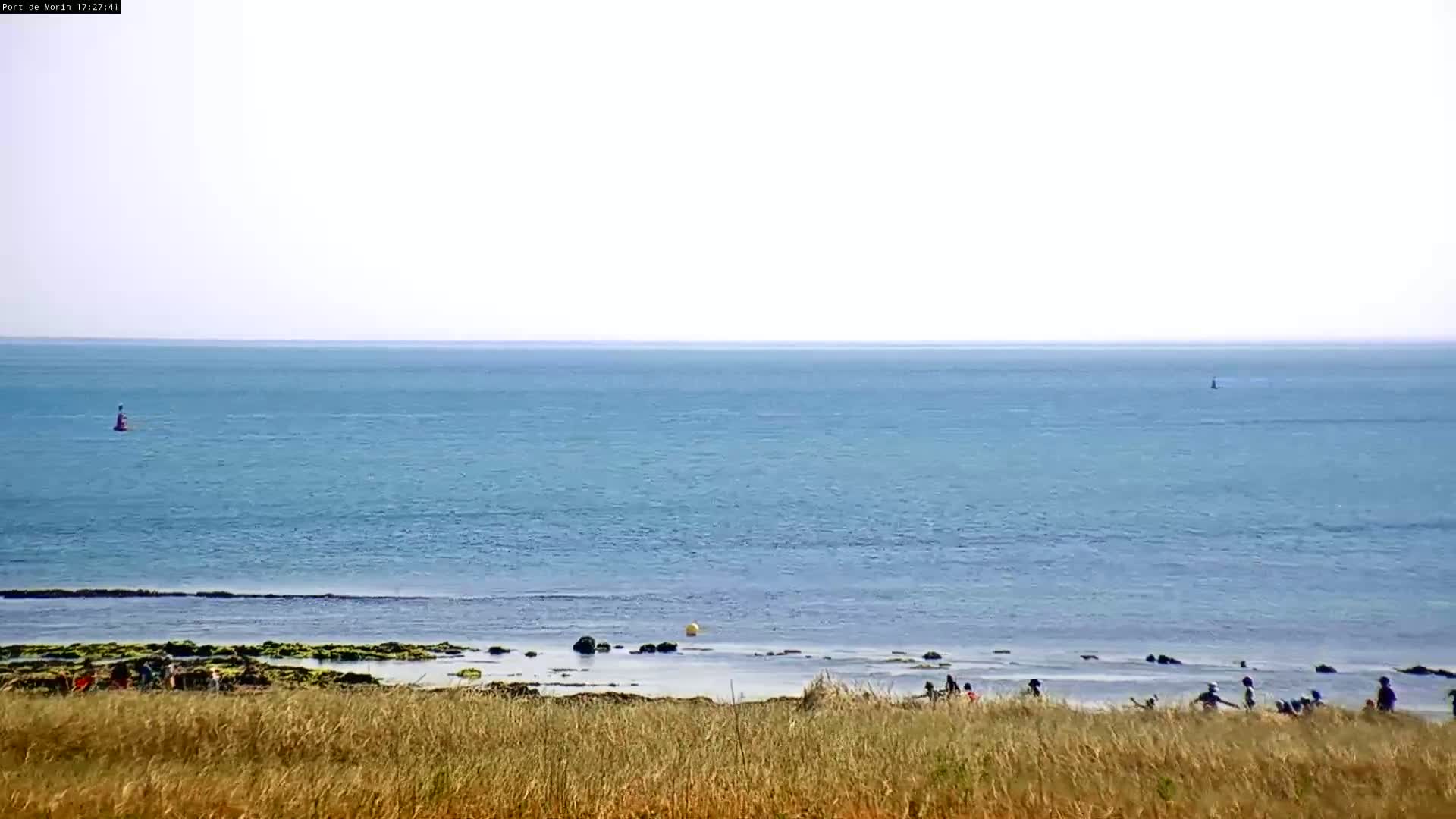 A calm, light-blue ocean stretches to the horizon under a bright, mostly clear sky, with a few people visible on a beach in the foreground, partially obscured by tall grass.