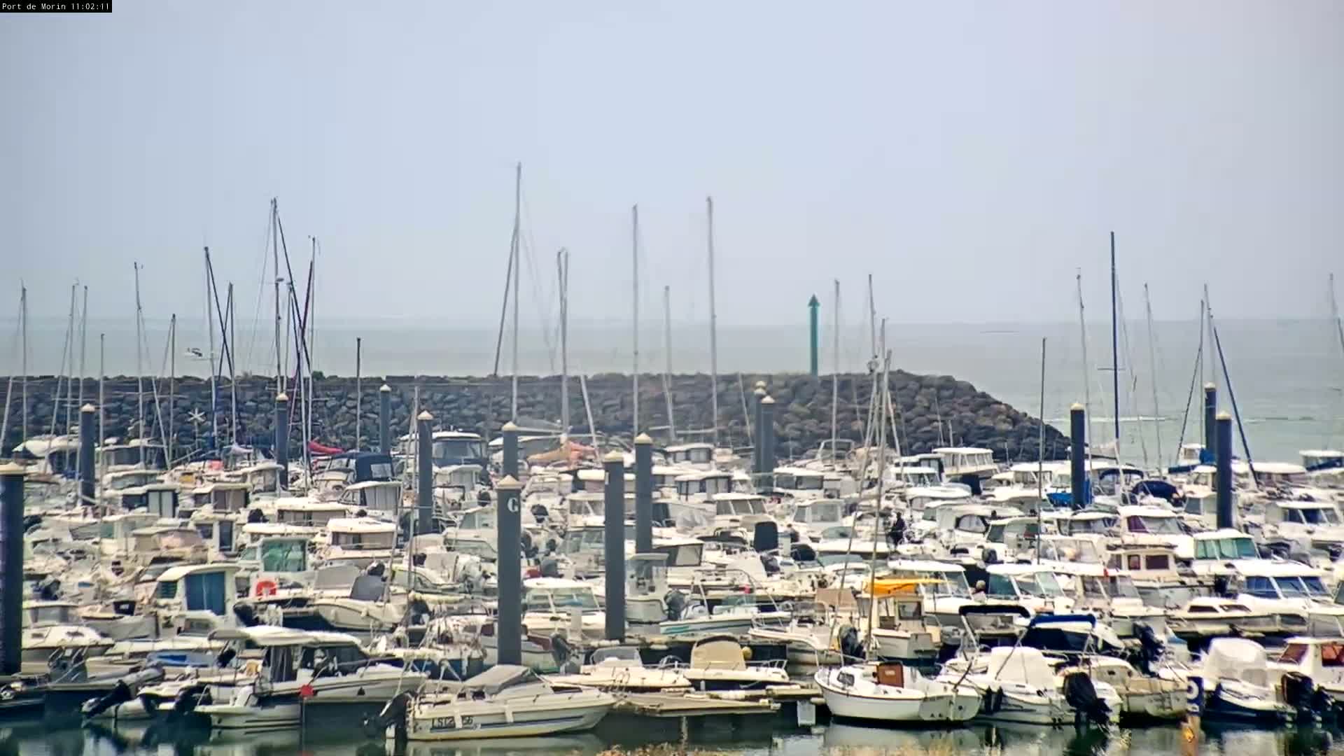 A marina filled with numerous boats and yachts is situated along a calm body of water under a hazy sky.