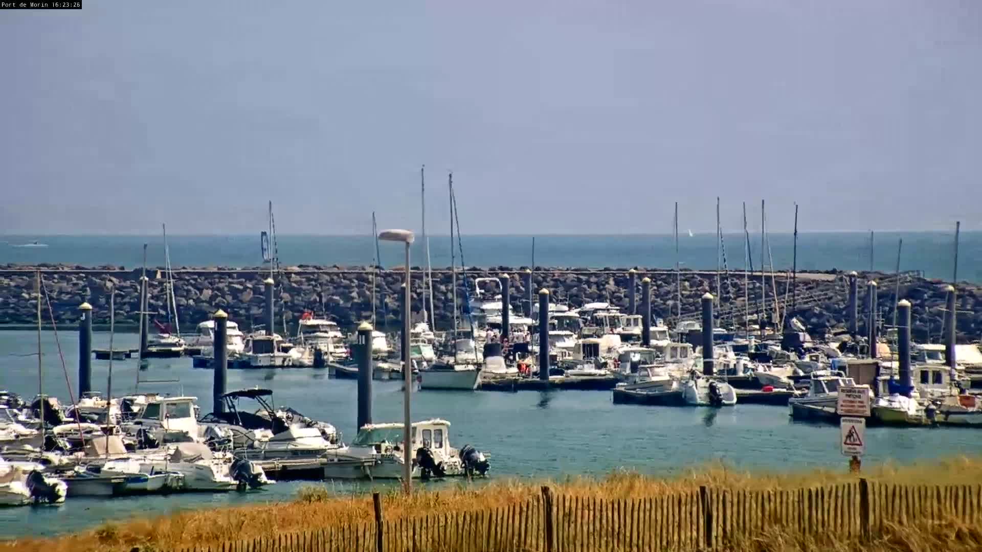 A marina filled with numerous boats is situated behind a low rock wall, under a mostly clear sky.
