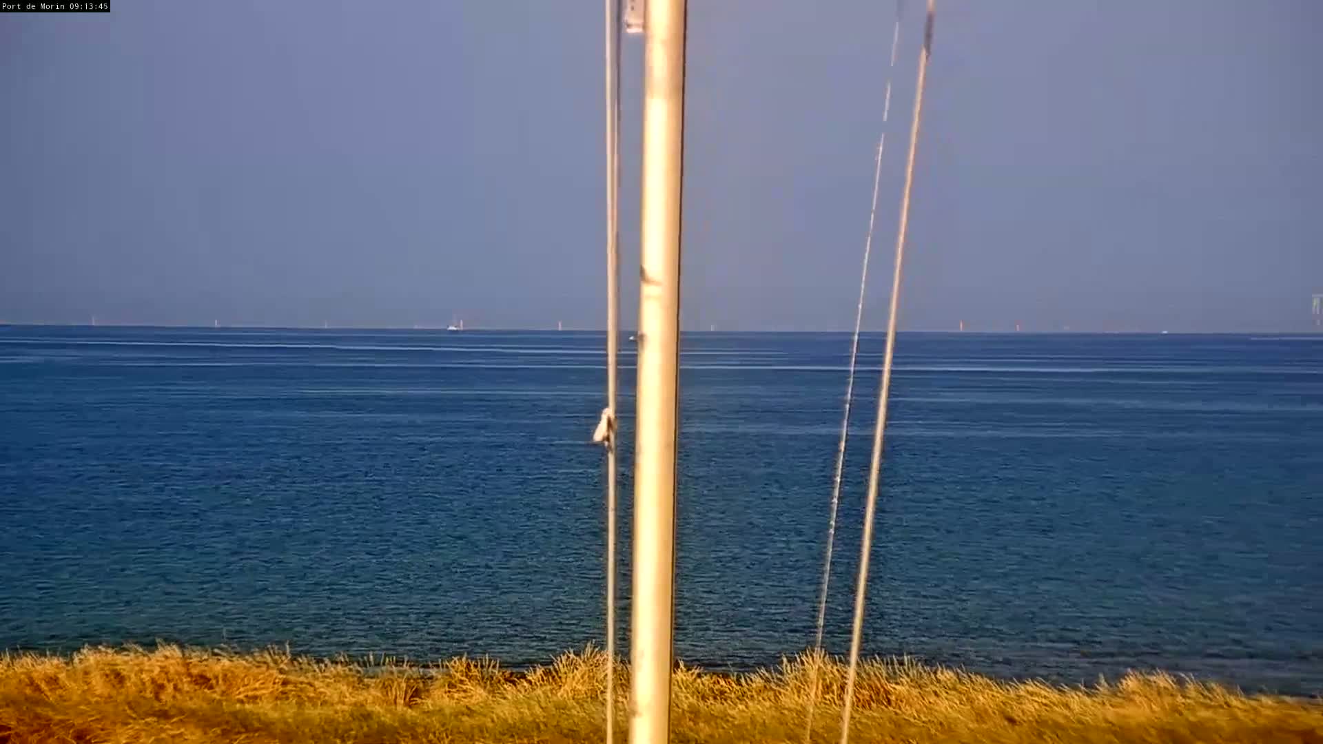 A calm, blue ocean stretches to the horizon, viewed from behind a tall pole with several lines attached, and a foreground of dry, tan grass.
