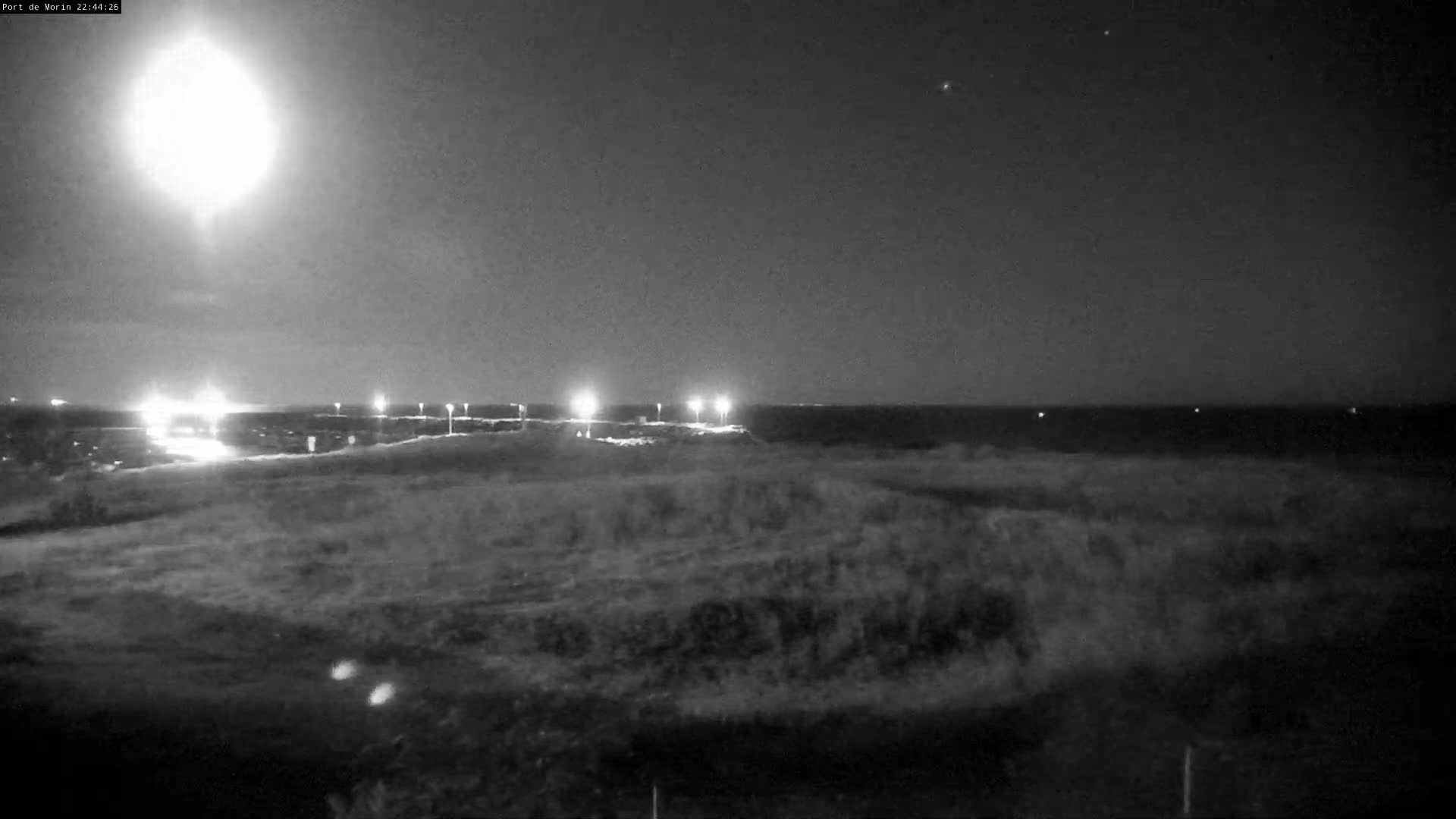 A nighttime, low-light image shows a bright light source in the sky above a dark landscape with distant lights and a body of water in the background.