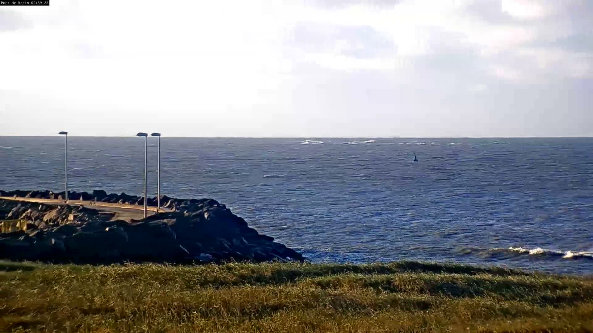 An overcast and windy day shows a rocky breakwater with a paved path and three streetlights extending into choppy dark blue ocean waters with whitecaps, seen from a grassy hillside.