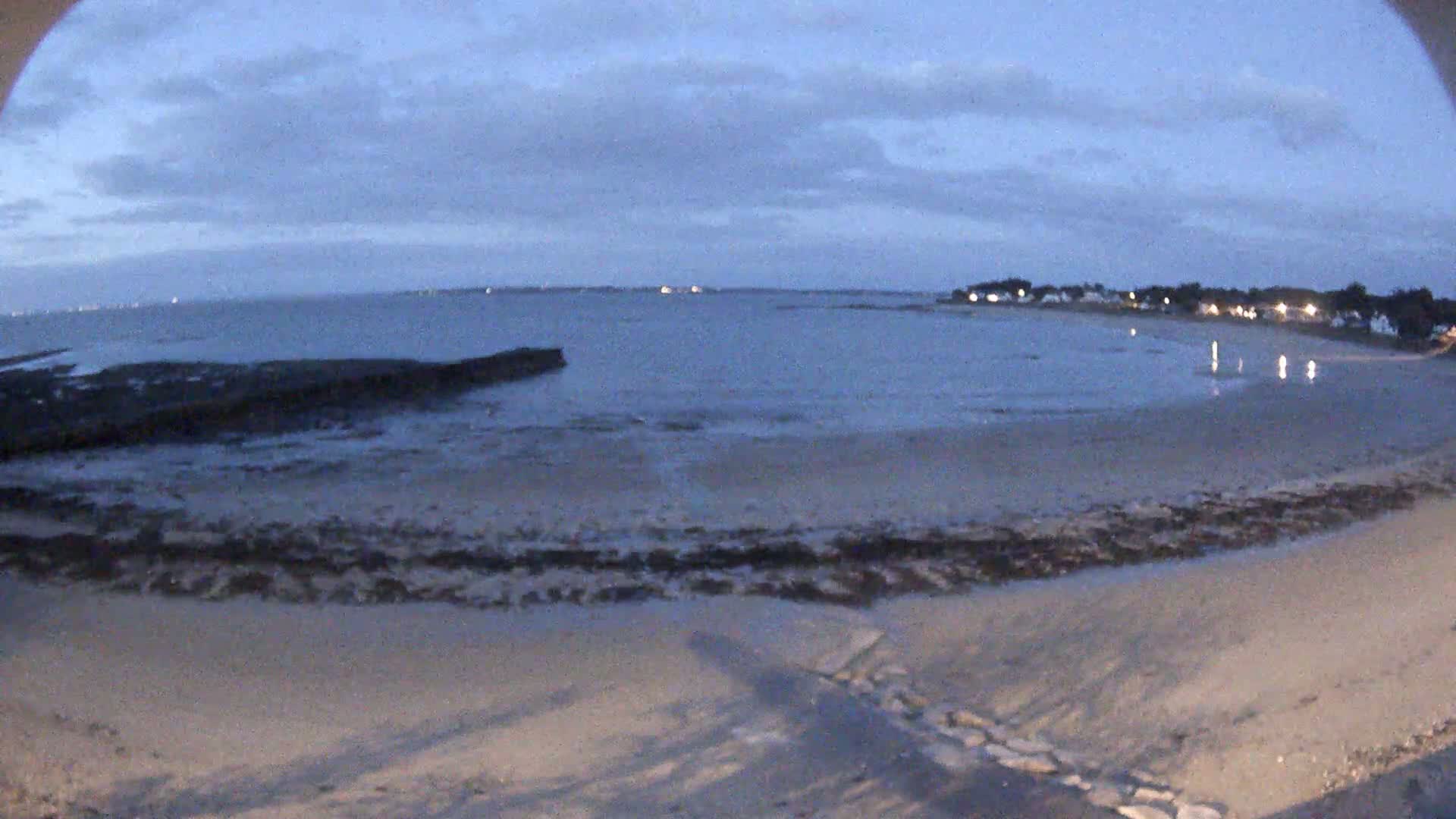 A sandy beach at low tide under a twilight sky, with calm water and a distant shoreline visible.