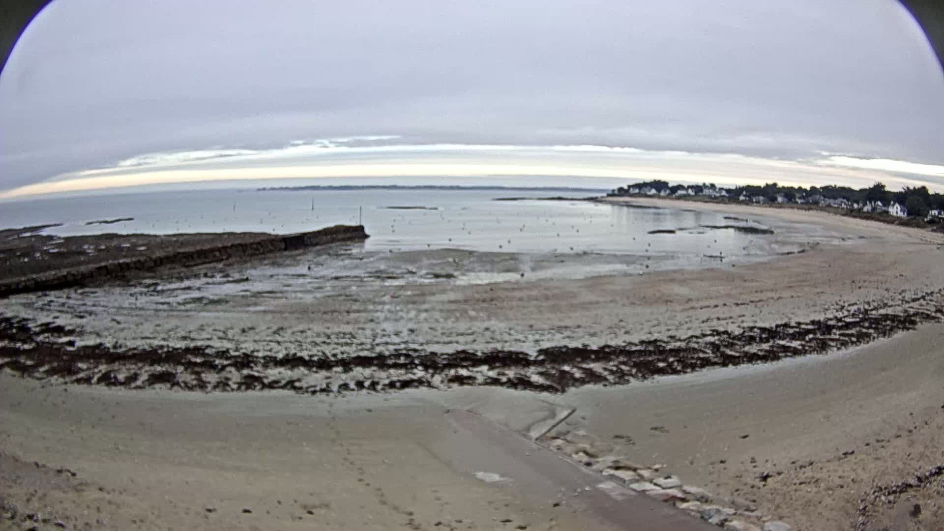 A wide, sandy beach at low tide stretches to a calm sea under a cloudy sky.