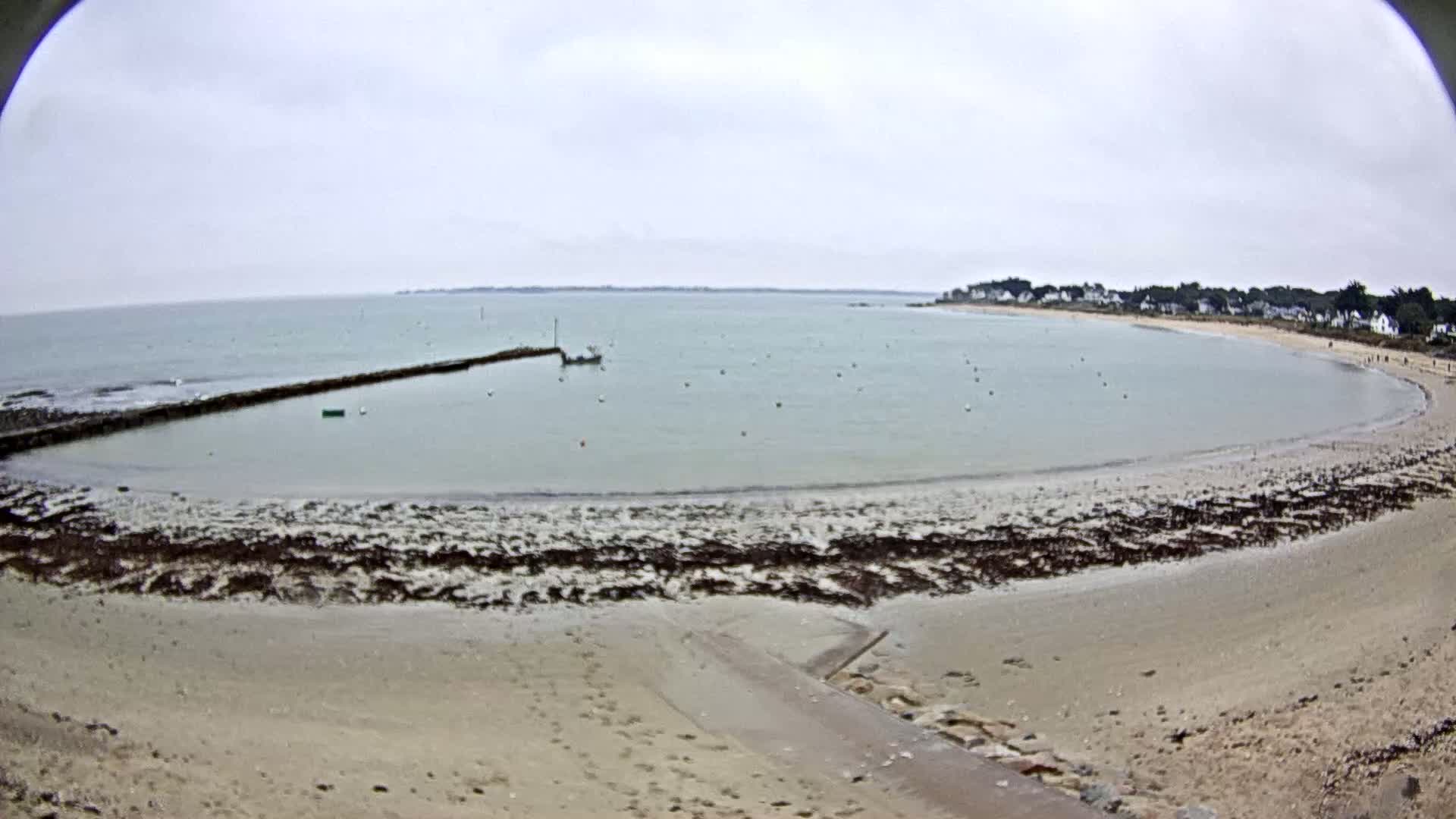 A sandy beach curves around a calm, light-green bay under an overcast sky, with a small boat and a low stone breakwater visible.