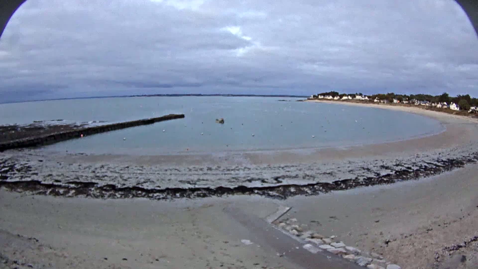A sandy beach curves around a calm bay under a cloudy sky, with a small pier and a few boats visible in the water.