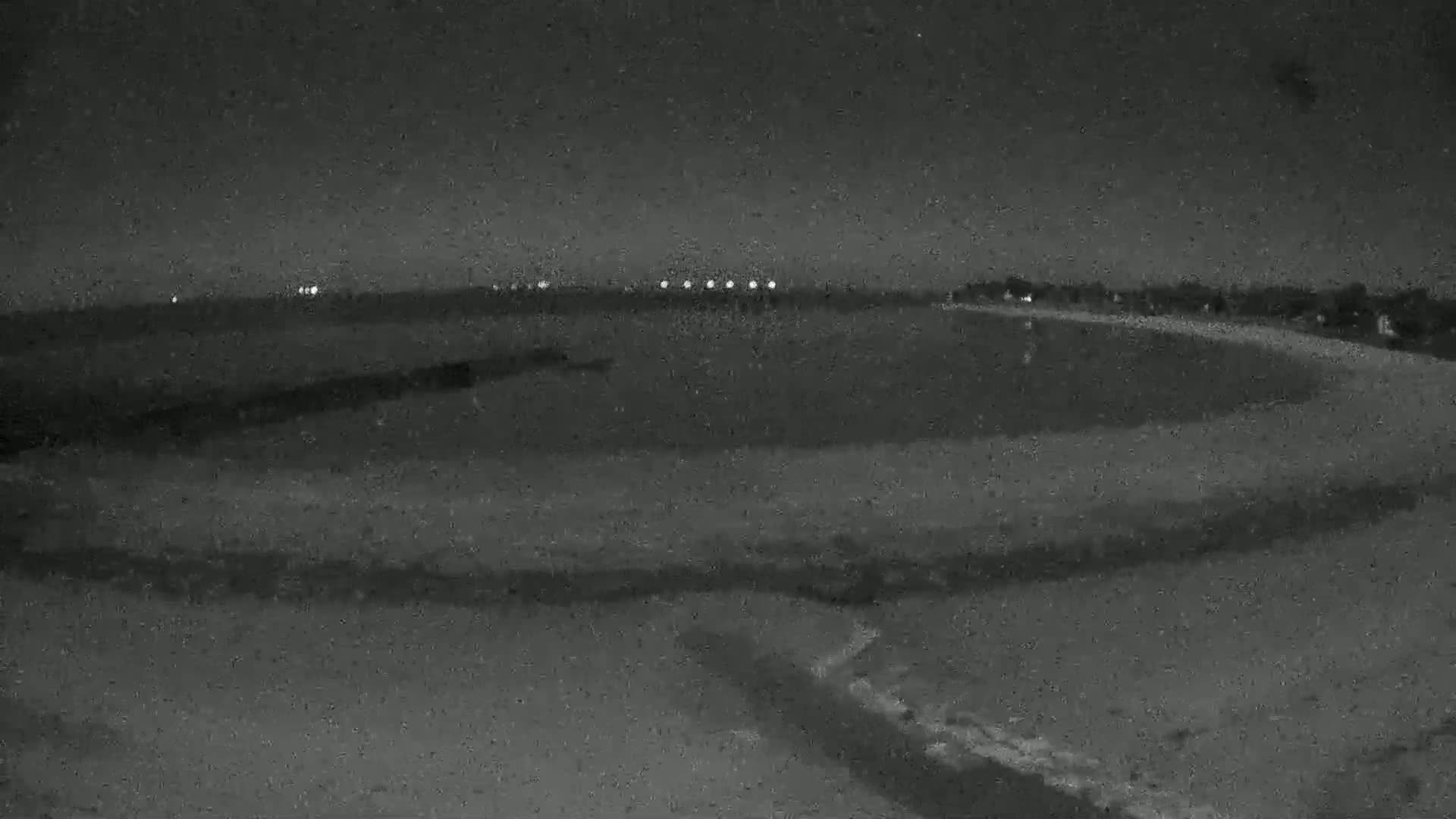 A dark, nighttime view of a flat, sandy area possibly a beach, extending to a distant horizon with a few faint lights visible.