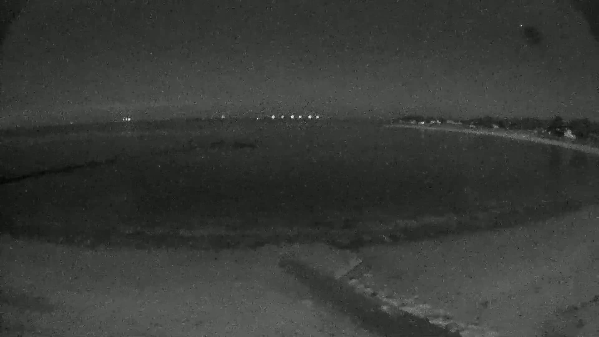 A nighttime, low-light image shows a dark, mostly flat expanse of land with a line of lights in the distance.