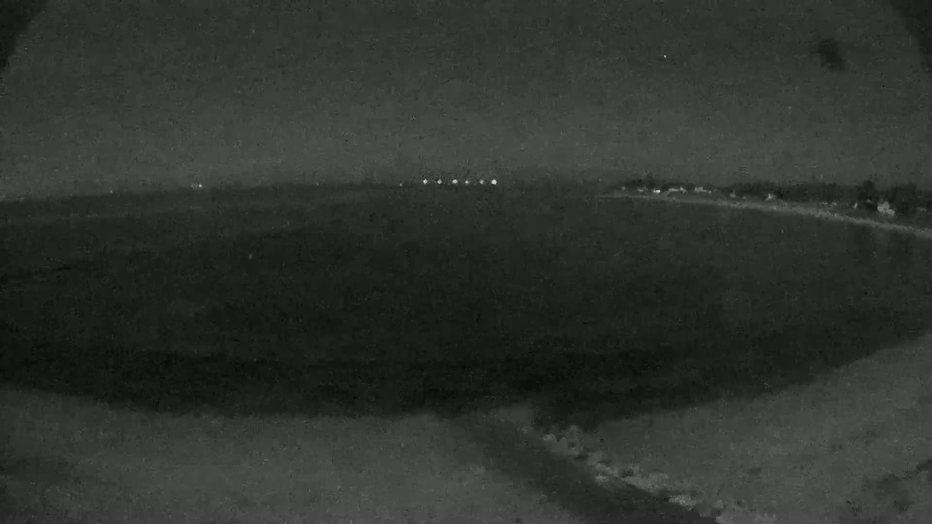 A nighttime, low-light view shows a dark expanse of land with a distant line of faint lights under a dark sky.
