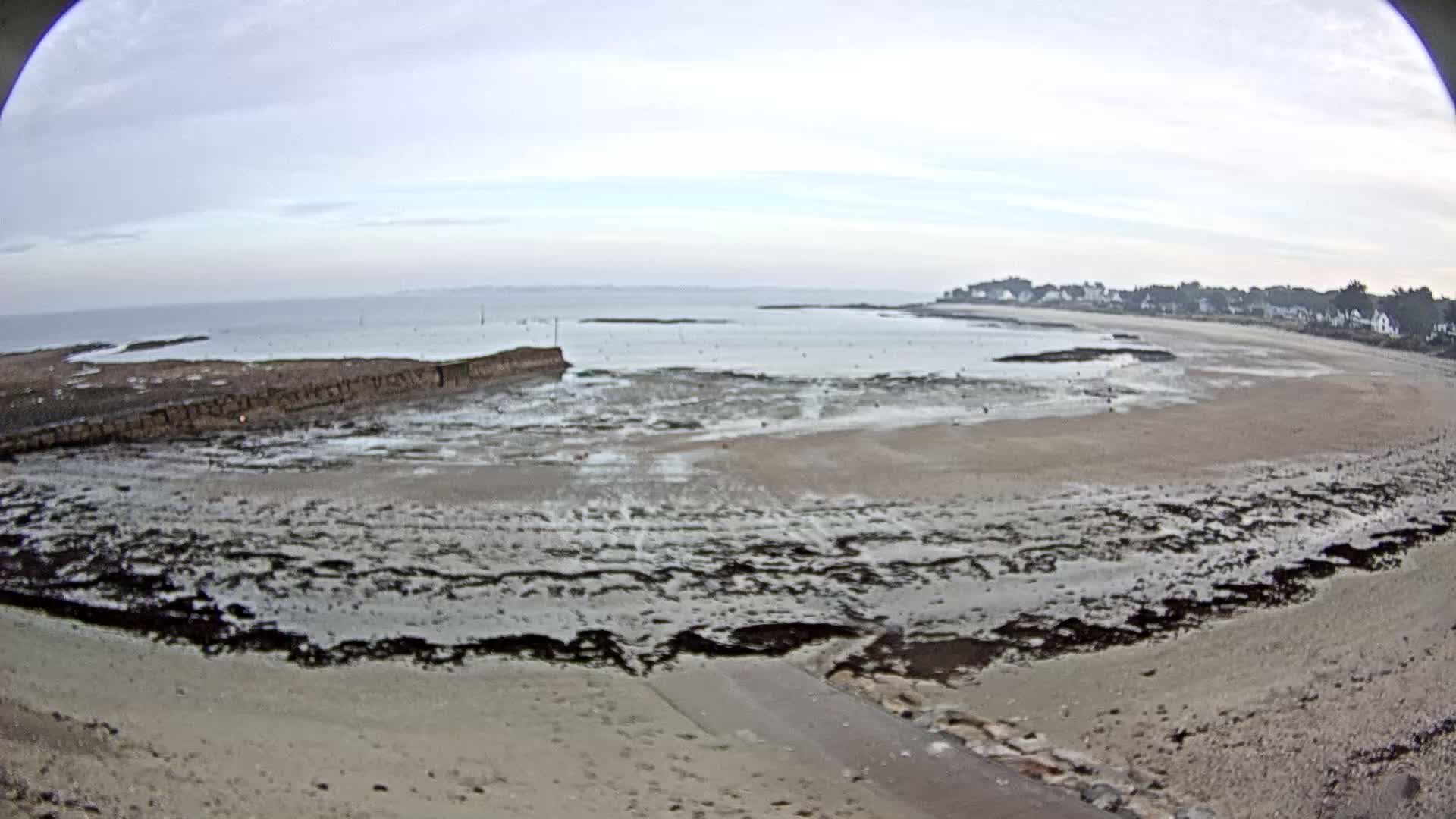 A wide, fisheye view shows a sandy beach at low tide, with dark, wet rocks and seaweed exposed, and a calm sea in the background under an overcast sky.