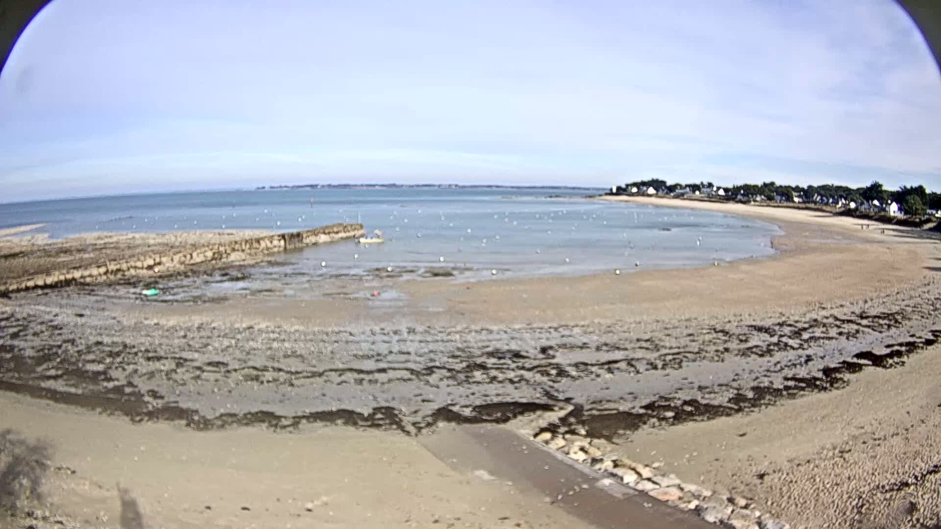 A sandy beach at low tide stretches towards a calm sea under a clear sky, with a small village visible in the distance.