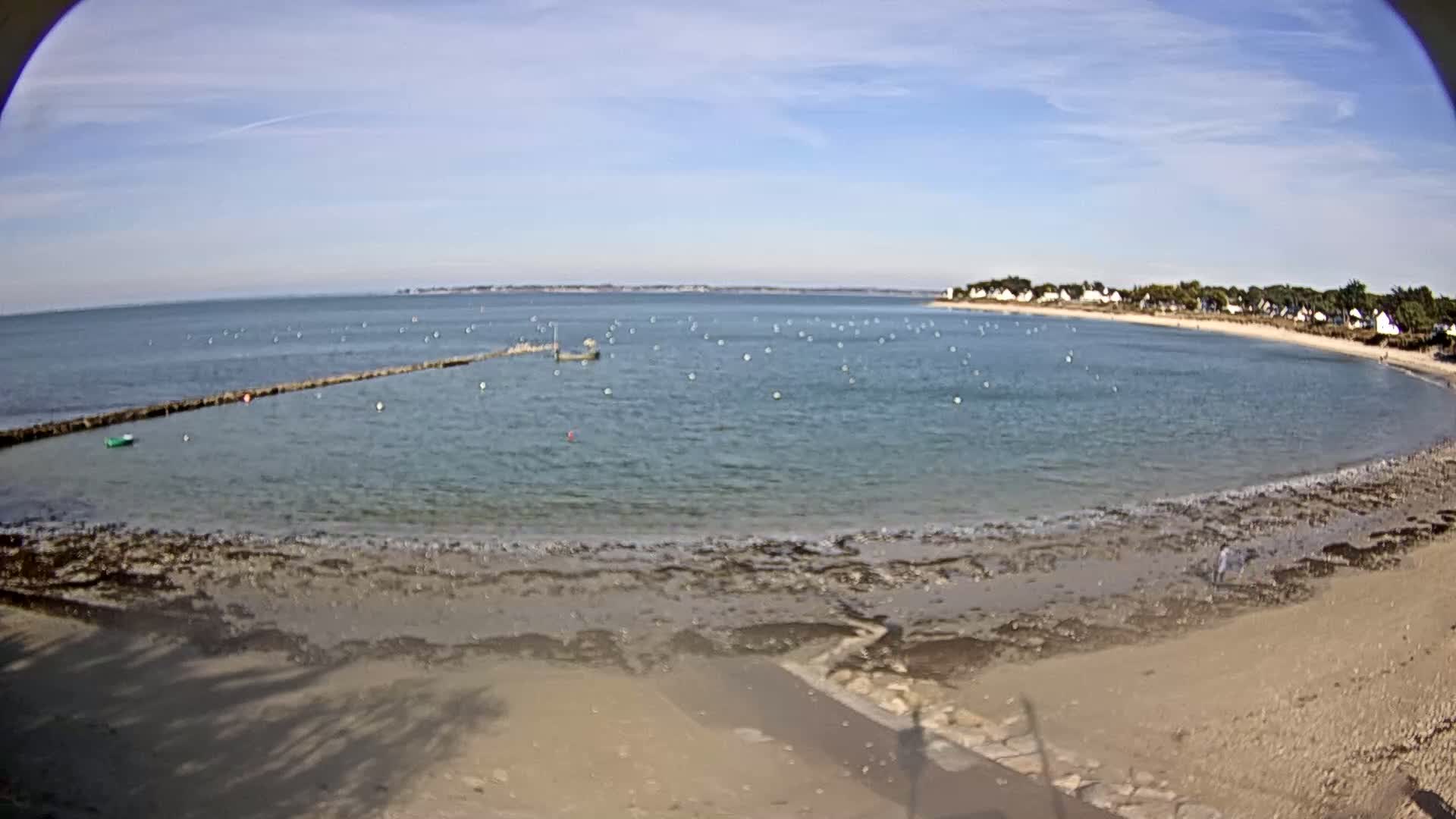 A calm, light-blue ocean bay with a sandy beach, a low stone barrier partially enclosing a swimming area, and a small cluster of houses visible on the far shore under a mostly sunny sky.