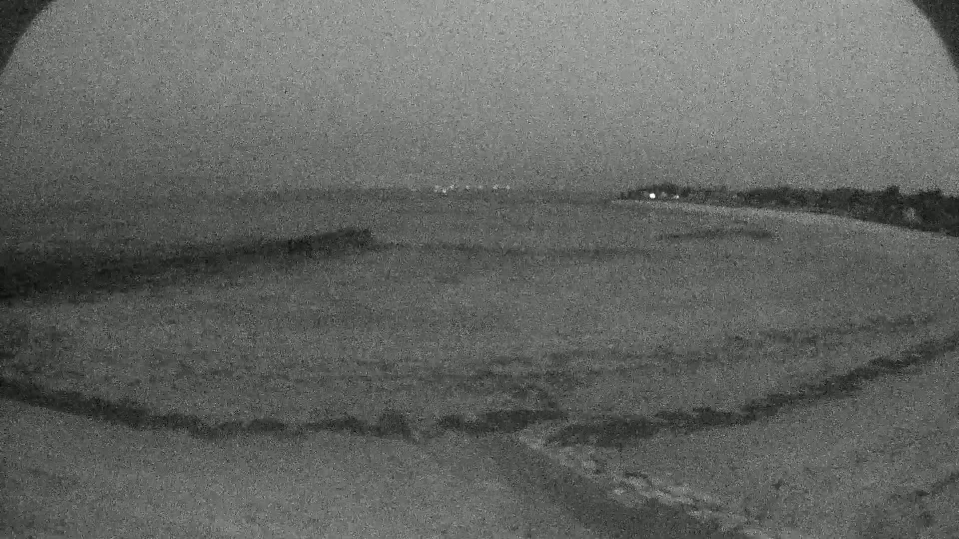 A low-light, grainy image shows a dark, possibly sandy beach at night, with calm water visible in the distance.