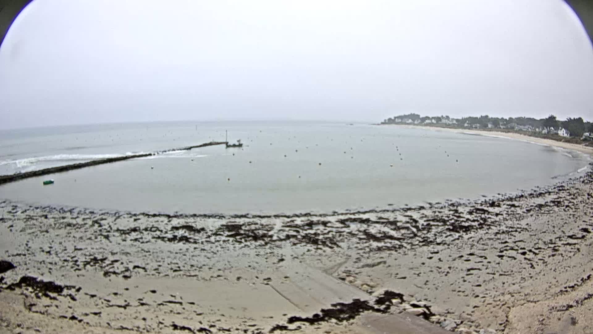 A calm, overcast day reveals a bay with a sandy beach, a small boat, and a rocky shoreline partially exposed at low tide.