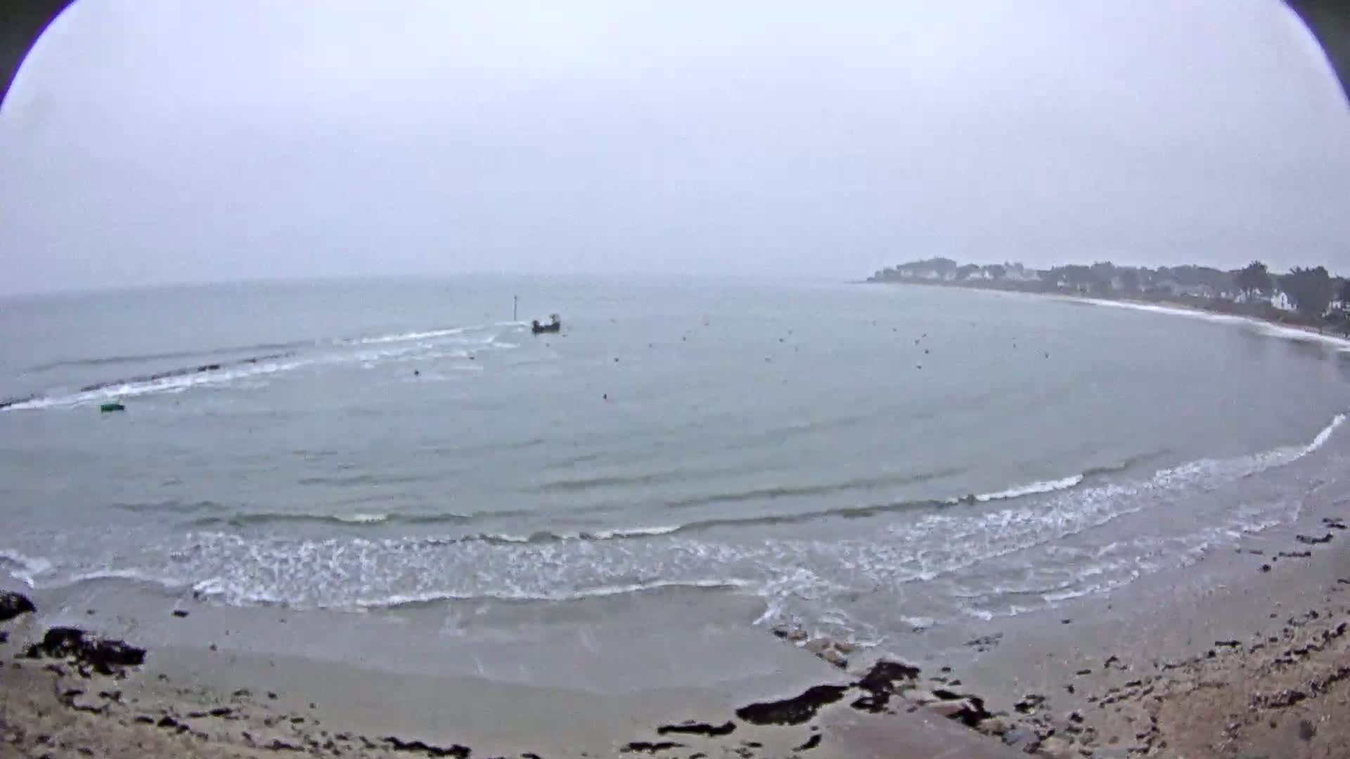 A small boat is visible on a grey, overcast day on a calm sea with small waves lapping a sandy beach.