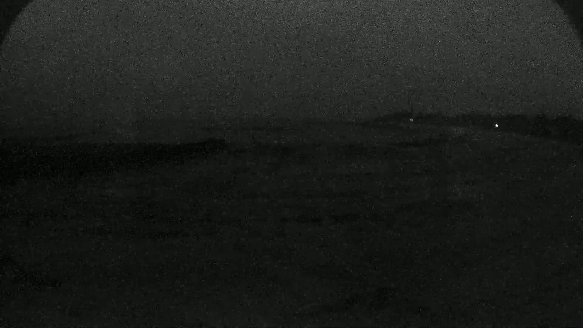 A dark, nighttime scene shows a flat, dark landscape with a few faint lights in the distance under a hazy sky.