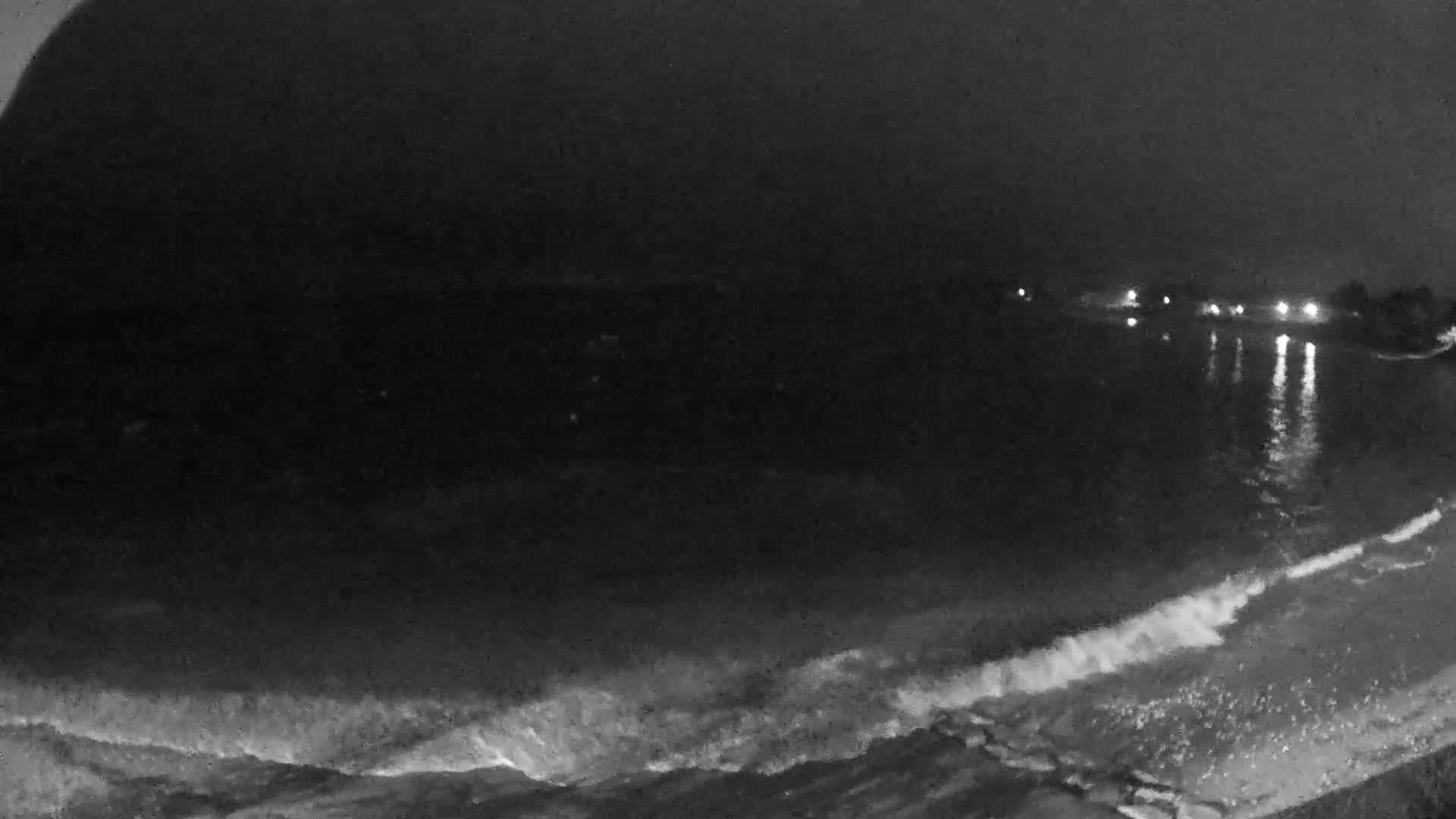 A nighttime, low-light view of gentle waves lapping a shoreline with distant lights reflecting on the water.